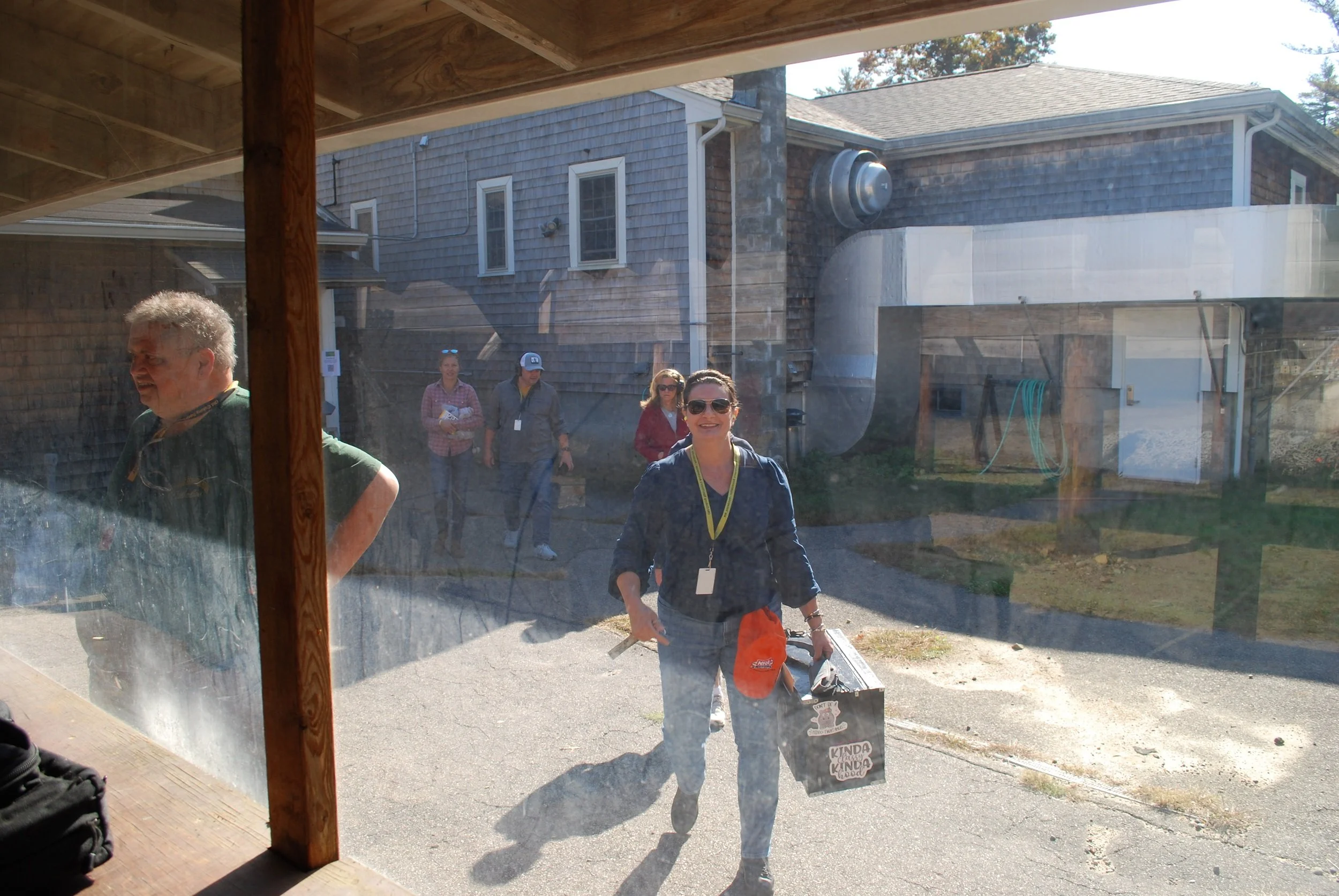 People walking outside view through a glass window, woman in sunglasses smiling and carrying a gift bag, some individuals in the background.