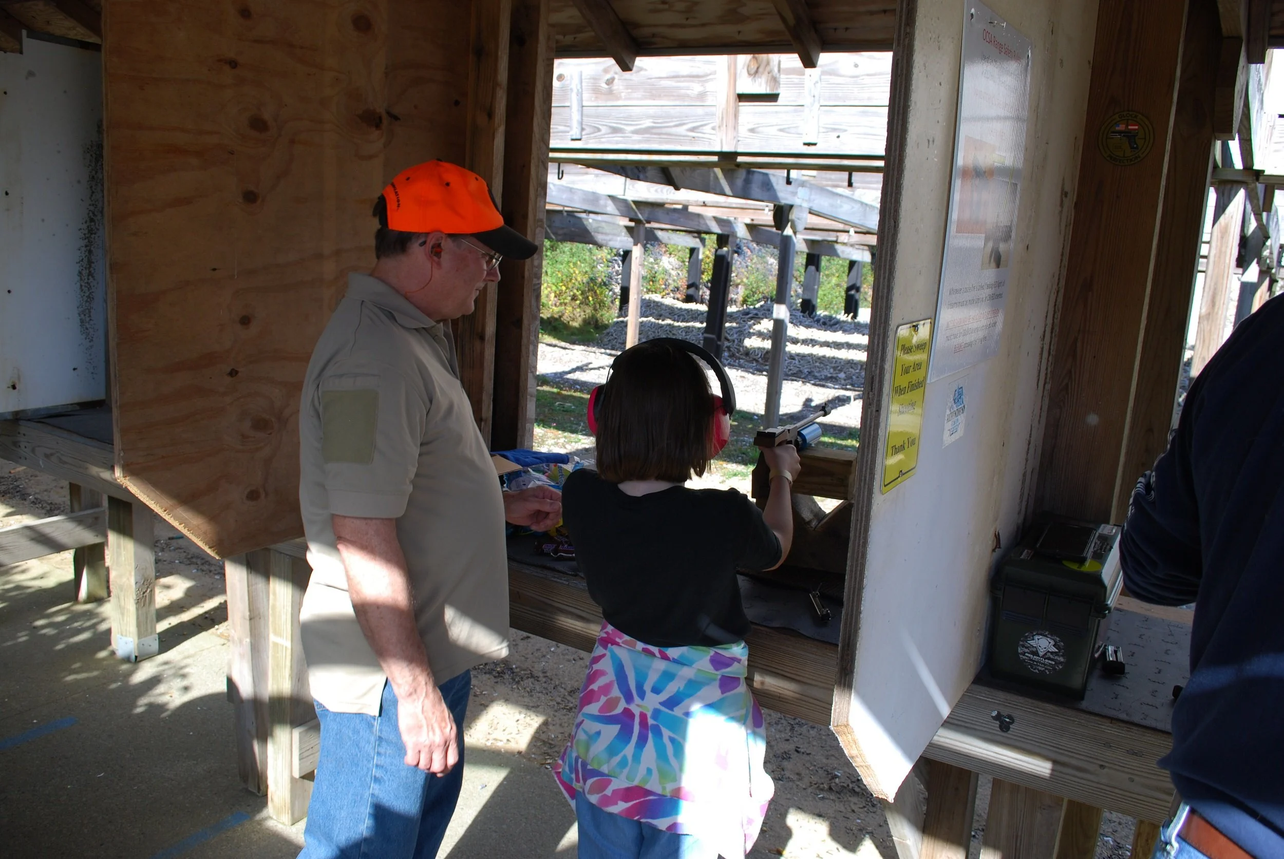 A young girl shooting a handgun at a shooting range with an instructor standing nearby in an outdoor setting.