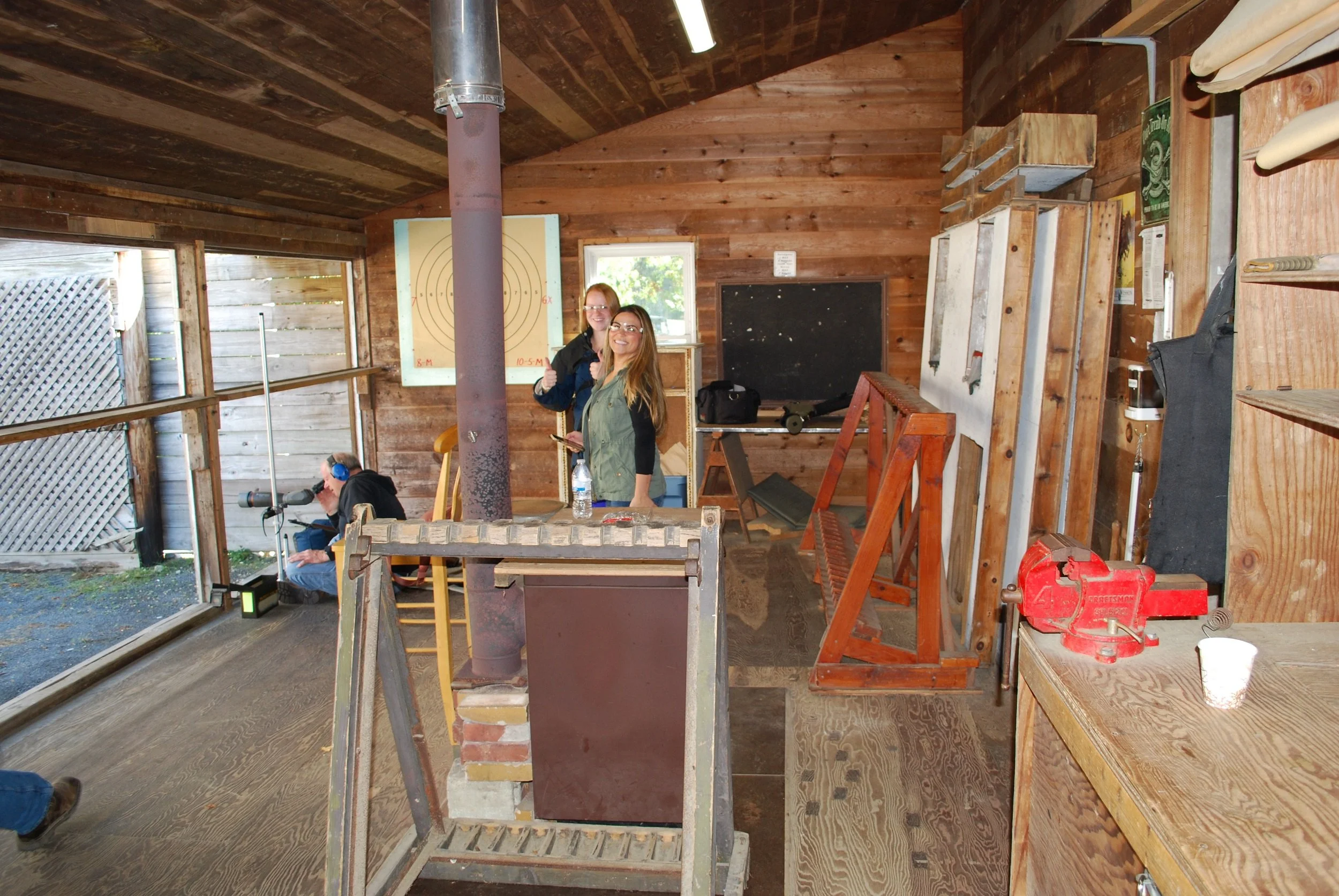Two smiling women standing inside a rustic wood workshop, giving a thumbs-up, with tools and woodworking supplies visible around them.