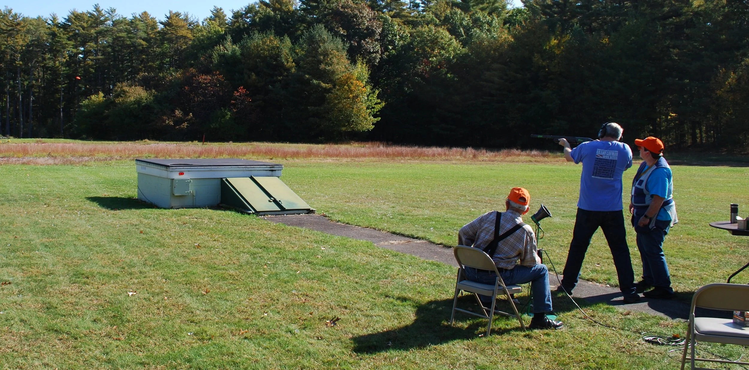Three people at a shooting range, one person aiming a rifle, two others observing, with trees in the background.