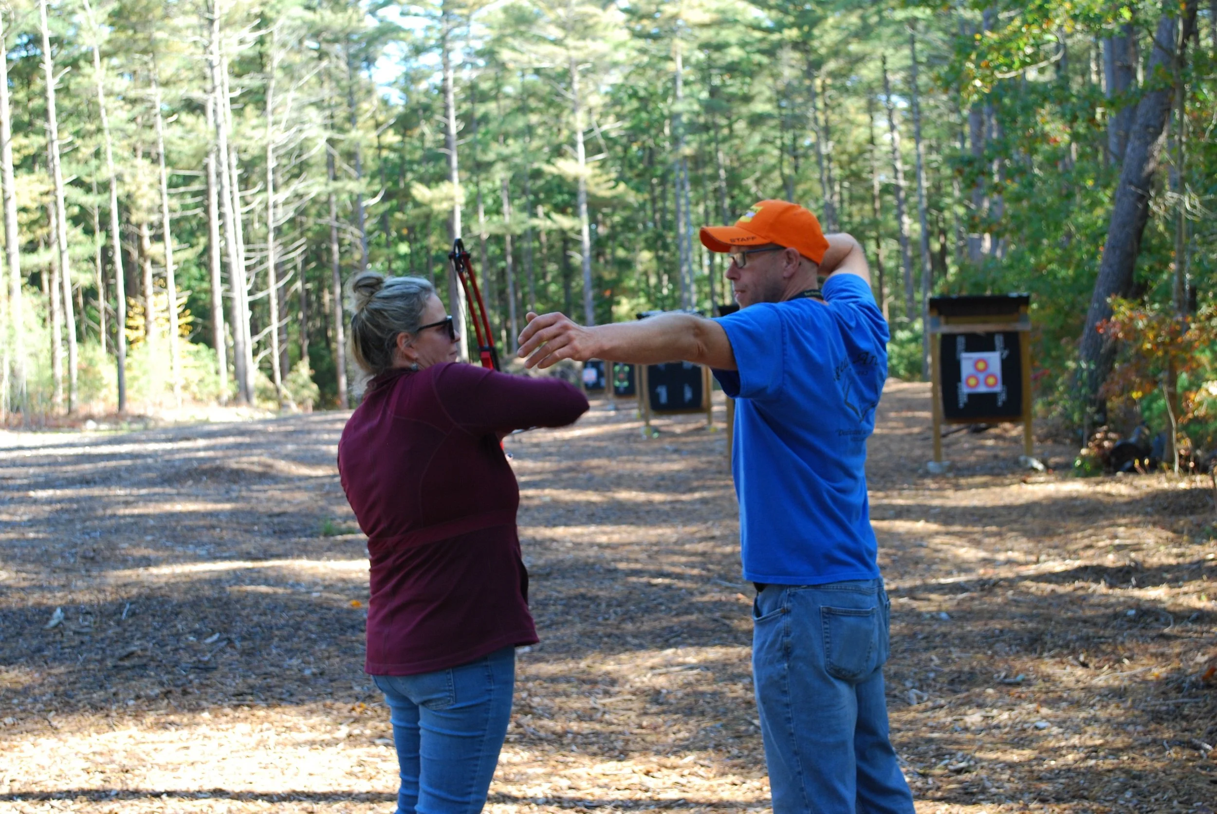 A woman with blonde hair tied in a bun and wearing sunglasses and a maroon jacket is holding a bow, and a man wearing sunglasses, an orange cap, a blue t-shirt, and jeans is instructing her on archery in a wooded outdoor area. There are archery targe
