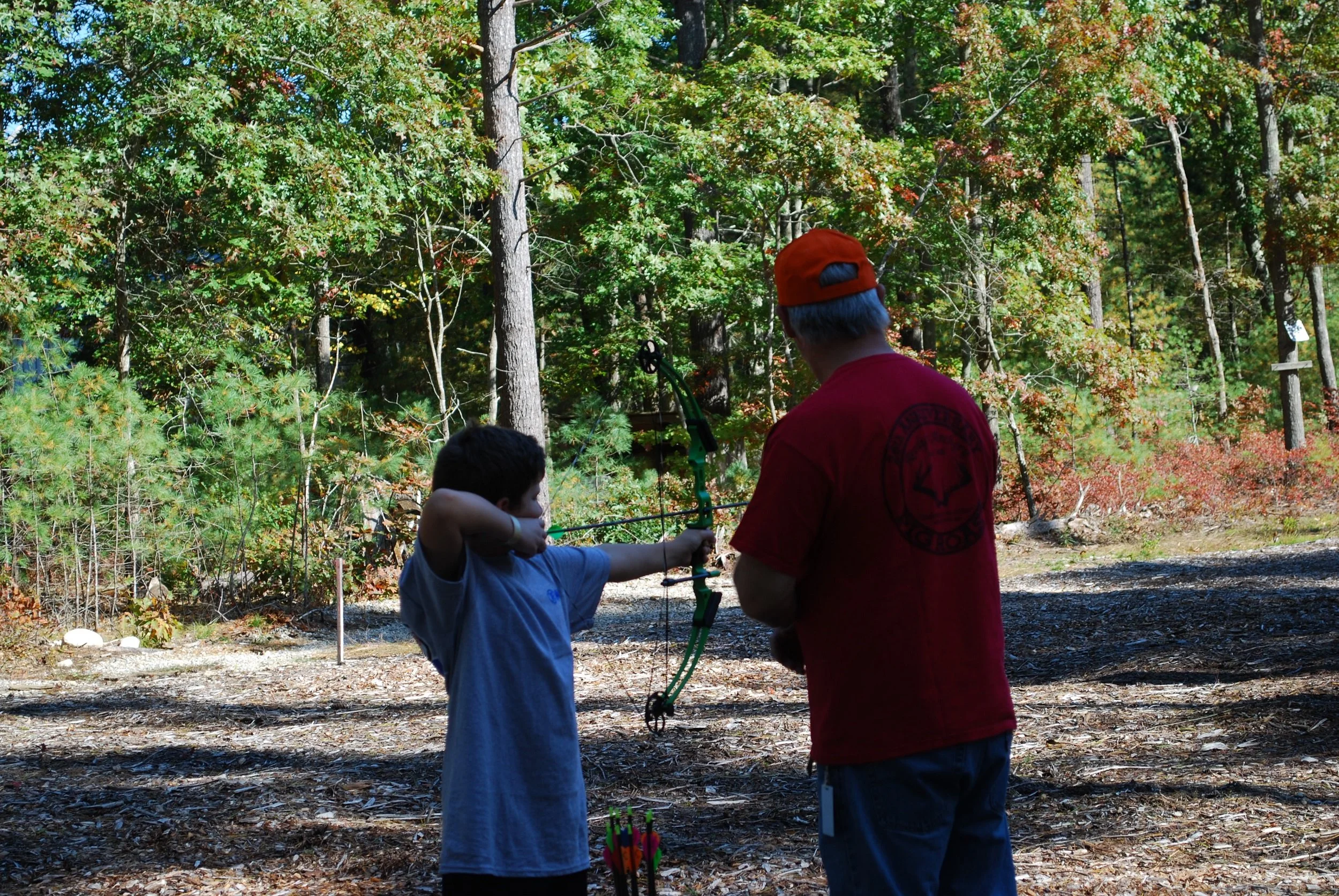A young boy in a blue shirt aiming a green archery bow with an adult man in a red shirt and orange cap observing in a wooded outdoor area.