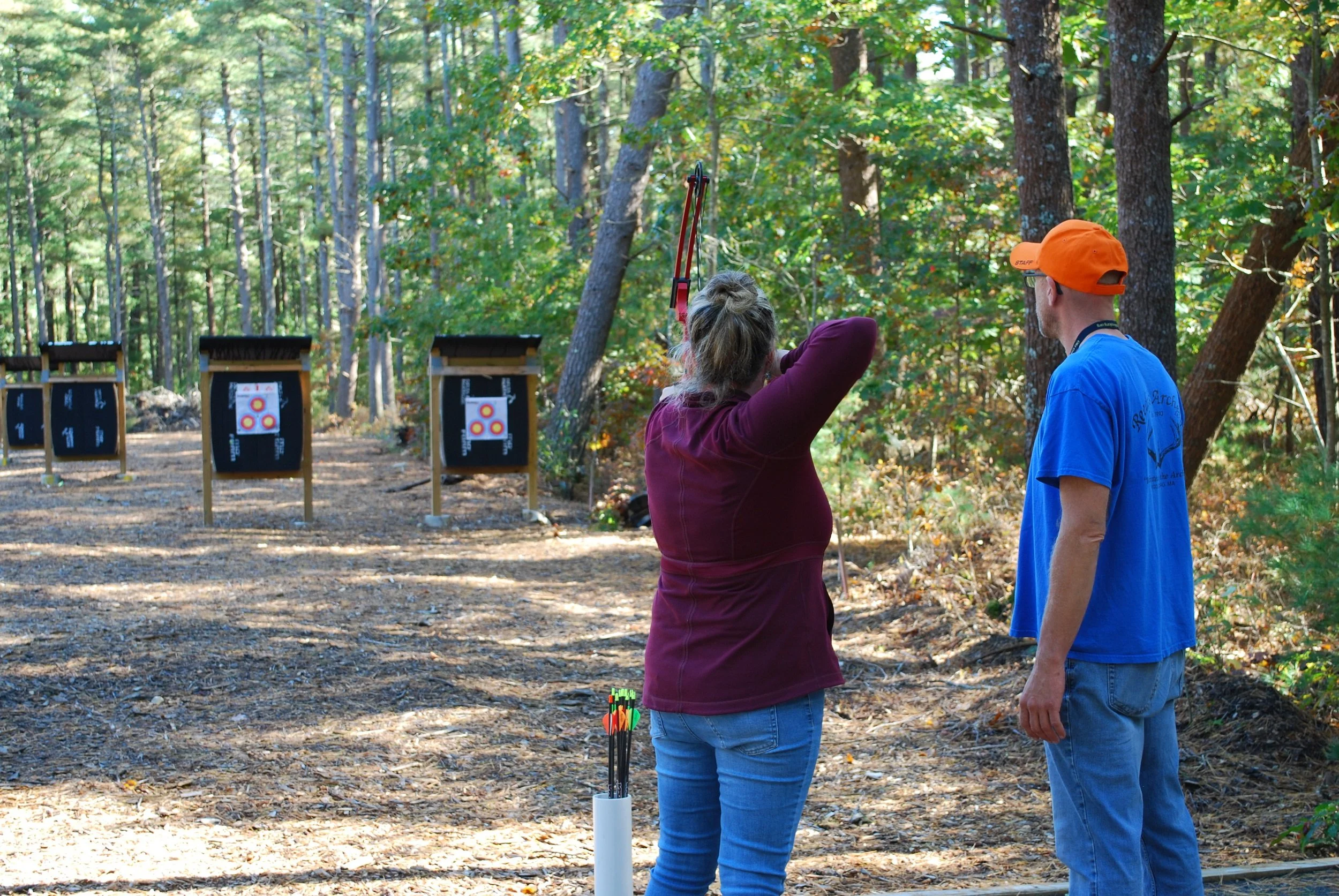 Two people at an outdoor archery range, with three targets in the background, surrounded by trees.