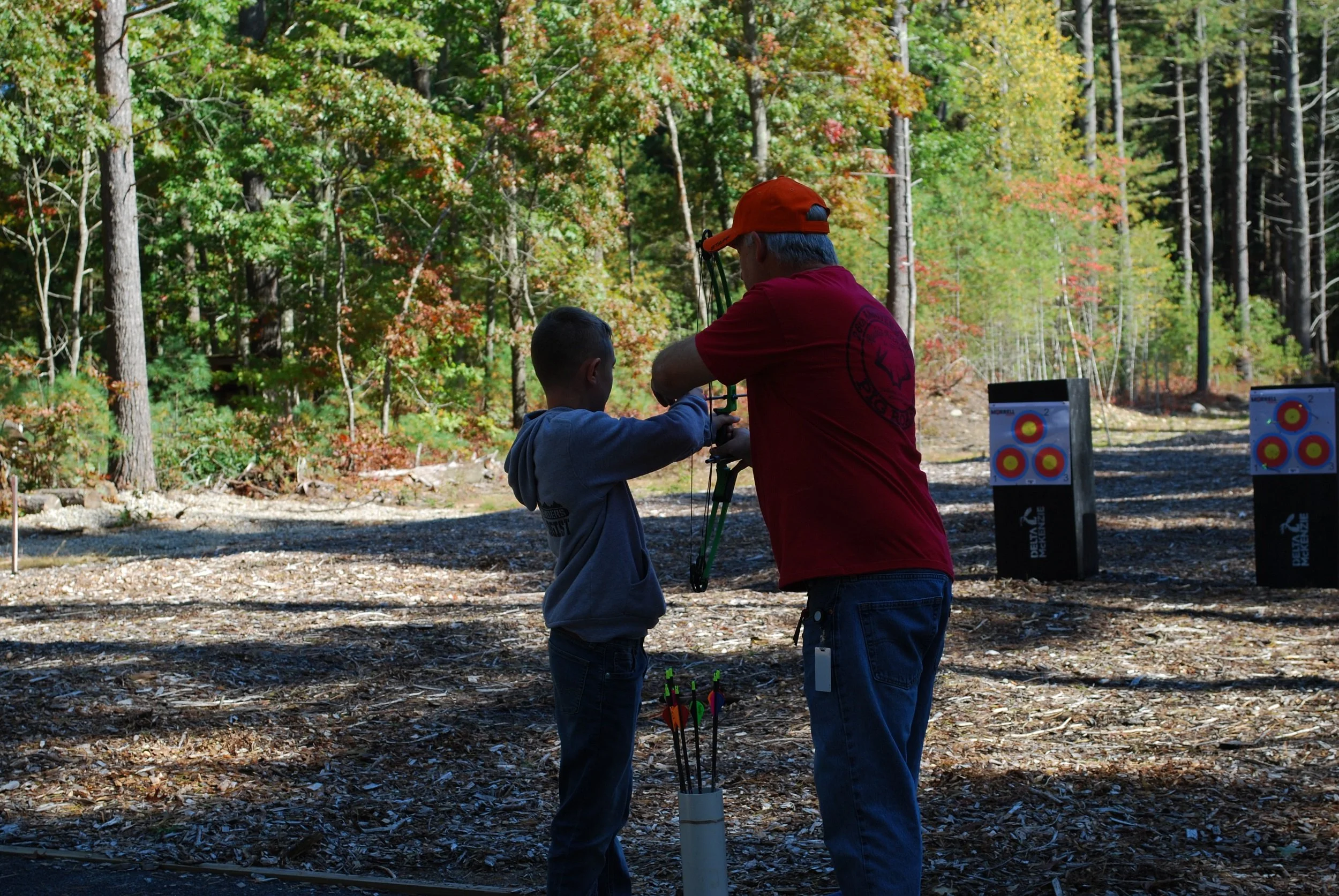 An adult teaching a child archery outdoors in a wooded area during autumn, with archery targets in the background.