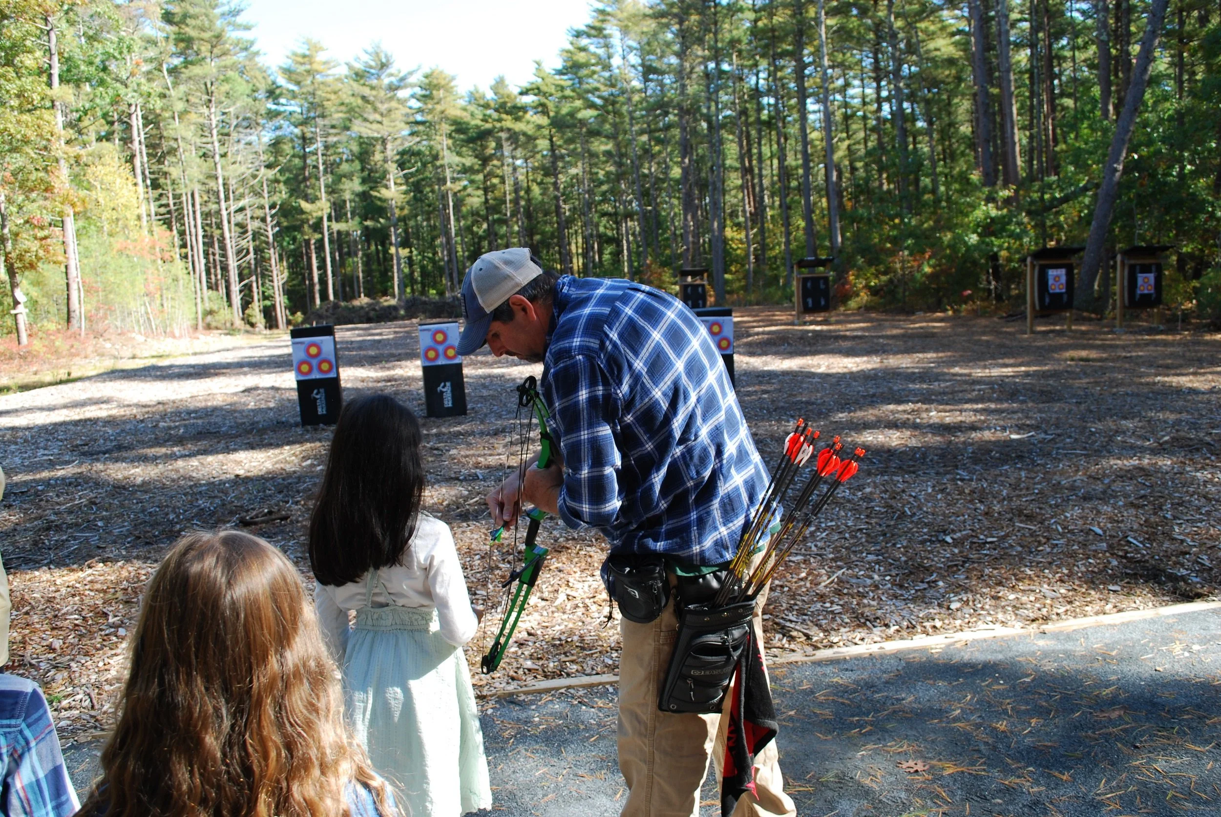 Man teaching children archery outdoors in a wooded area, with target boards in the background.