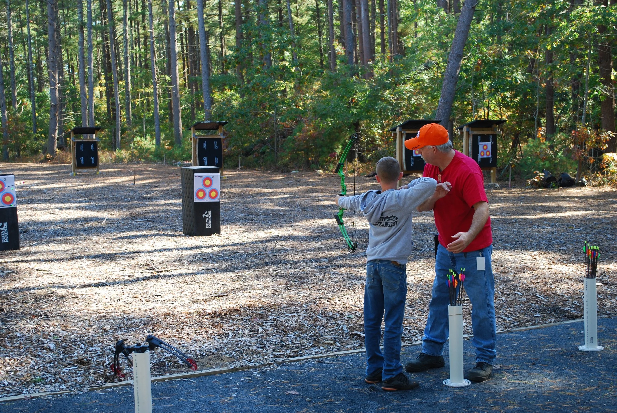 An outdoor archery range with targets set up in a wooded area. A boy is receiving instruction from an adult man, both preparing to shoot arrows.