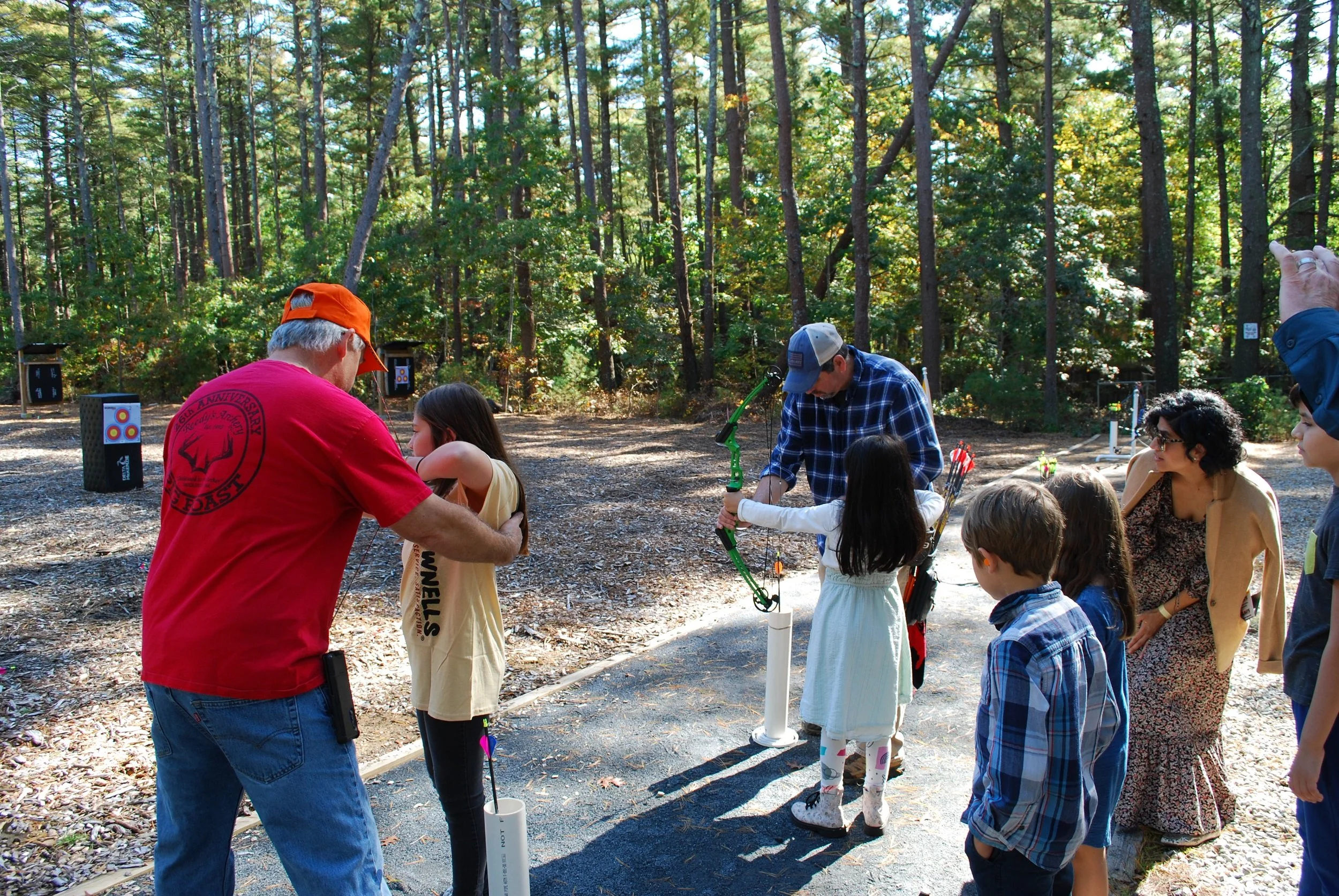 Group of children and adults at an outdoor archery activity in a forested area, with targets set up in the background.