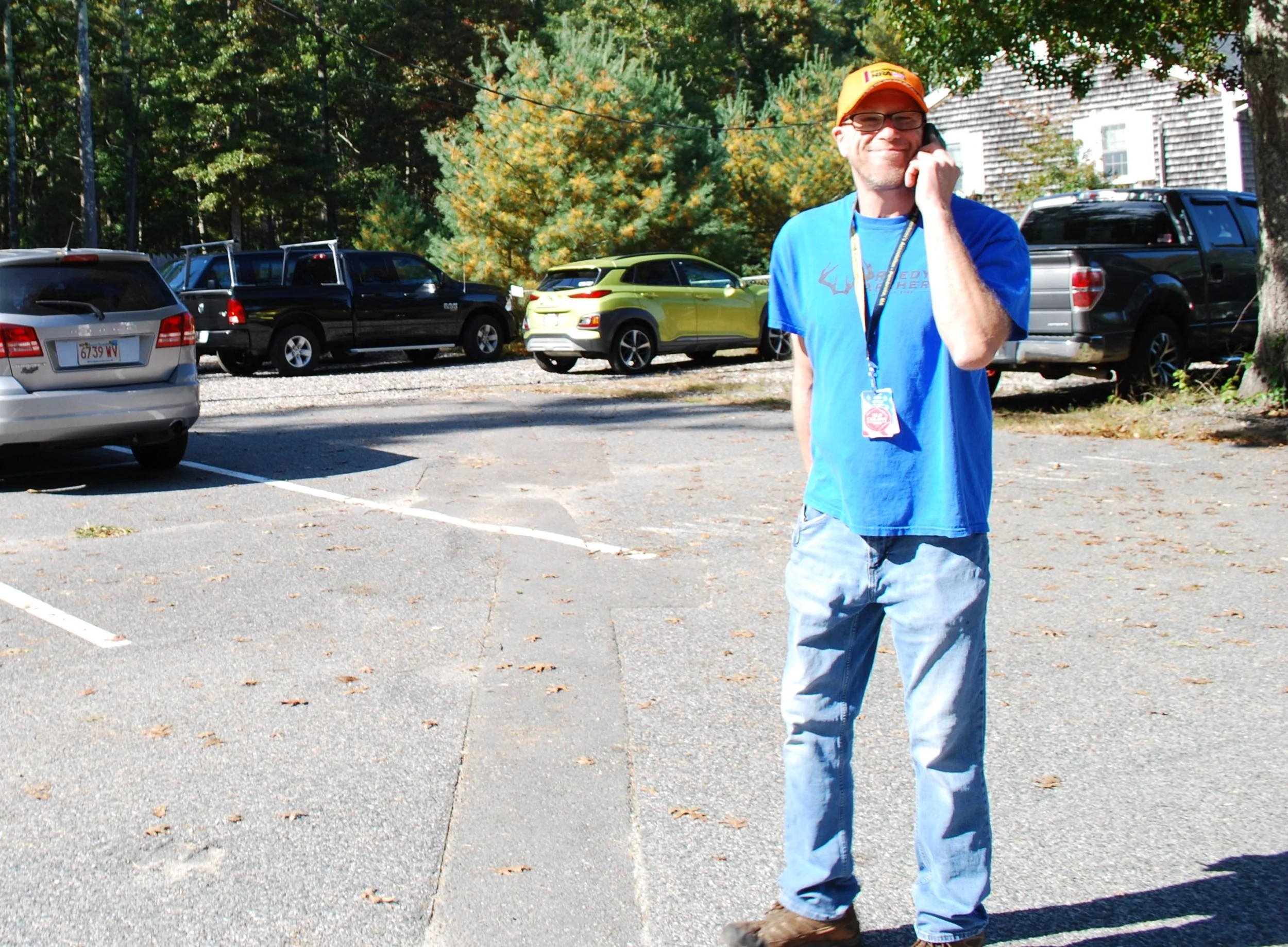 A man wearing a blue t-shirt, jeans, a yellow cap, and glasses is standing in a parking lot, talking on a cellphone, with cars parked behind him and trees in the background.
