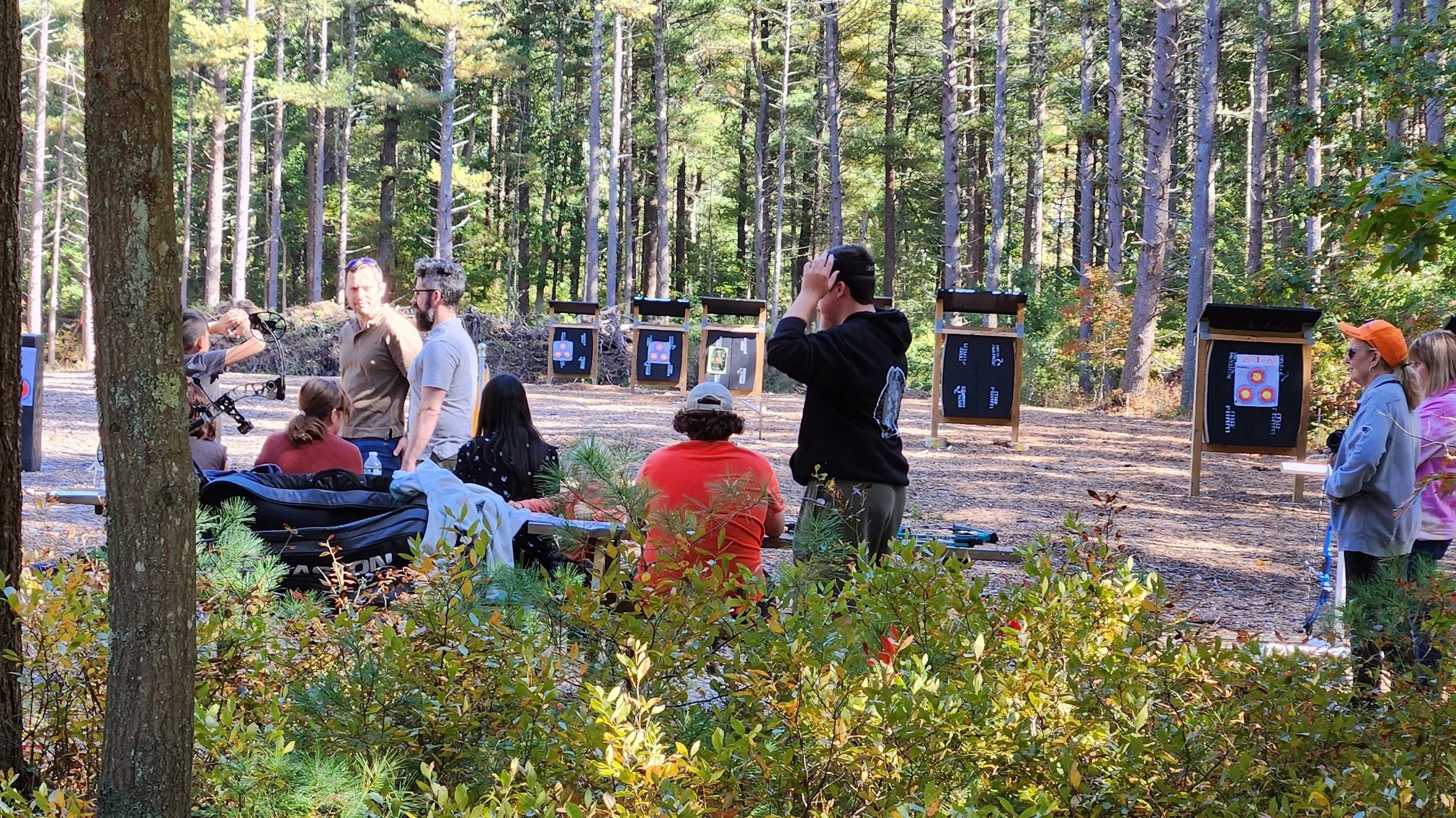 People participating in an outdoor archery activity in a wooded area with targets set up in the background. Some are seated at a table, others are standing and preparing to shoot.