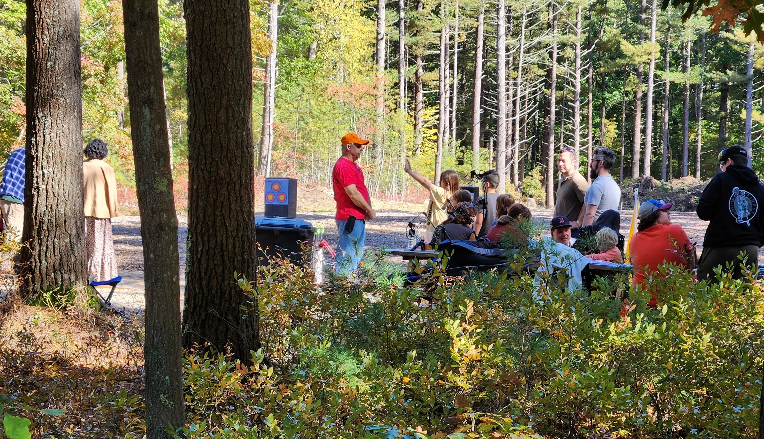 People gathered outdoors in a forest during daytime for archery activity or event, with some sitting on benches and others standing, surrounded by tall trees and autumn foliage.