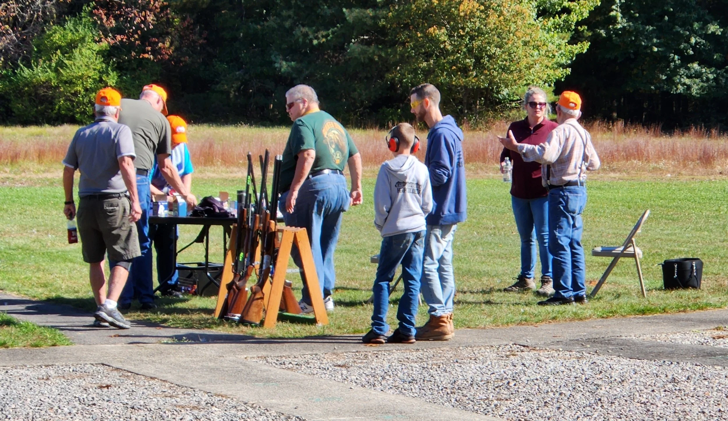 Group of people outdoors at a shooting range, with rifles on display and some wearing safety ear and eye protection.