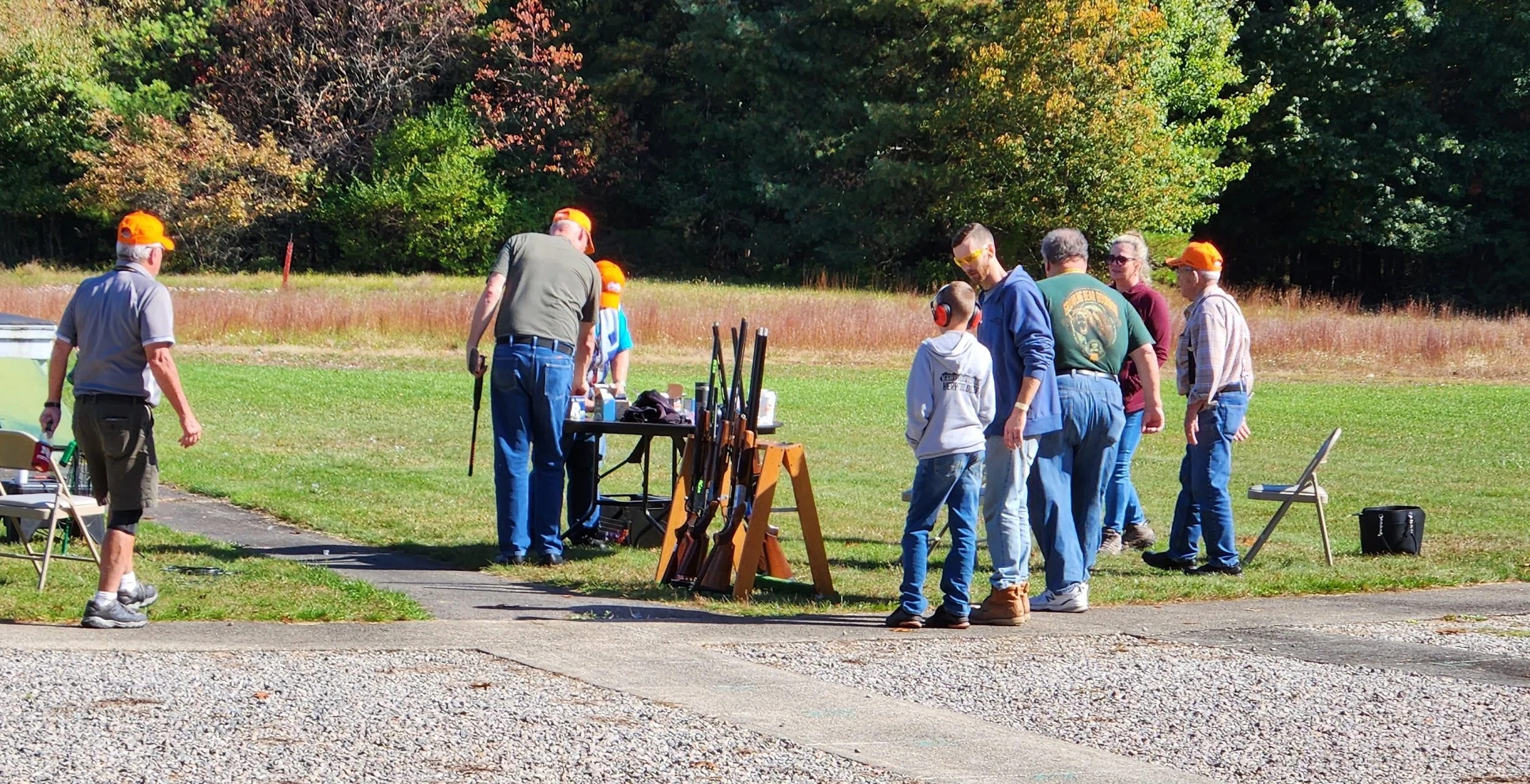Group of people at an outdoor shooting range with rifles and safety gear, set in a grassy area surrounded by trees.