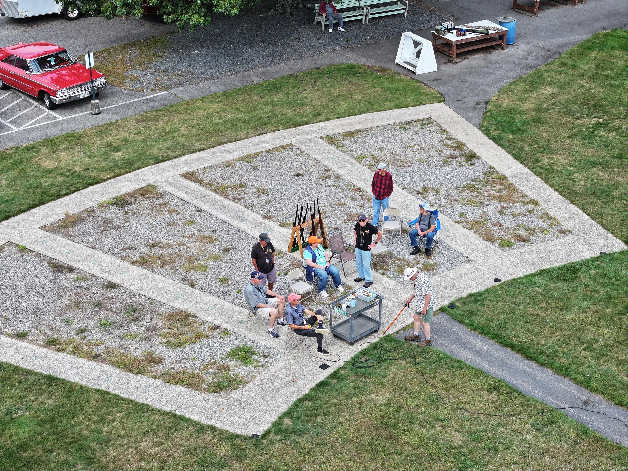 A group of people gathered on a trap field with chairs, easels, and supplies, Some are sitting, and one man is standing.