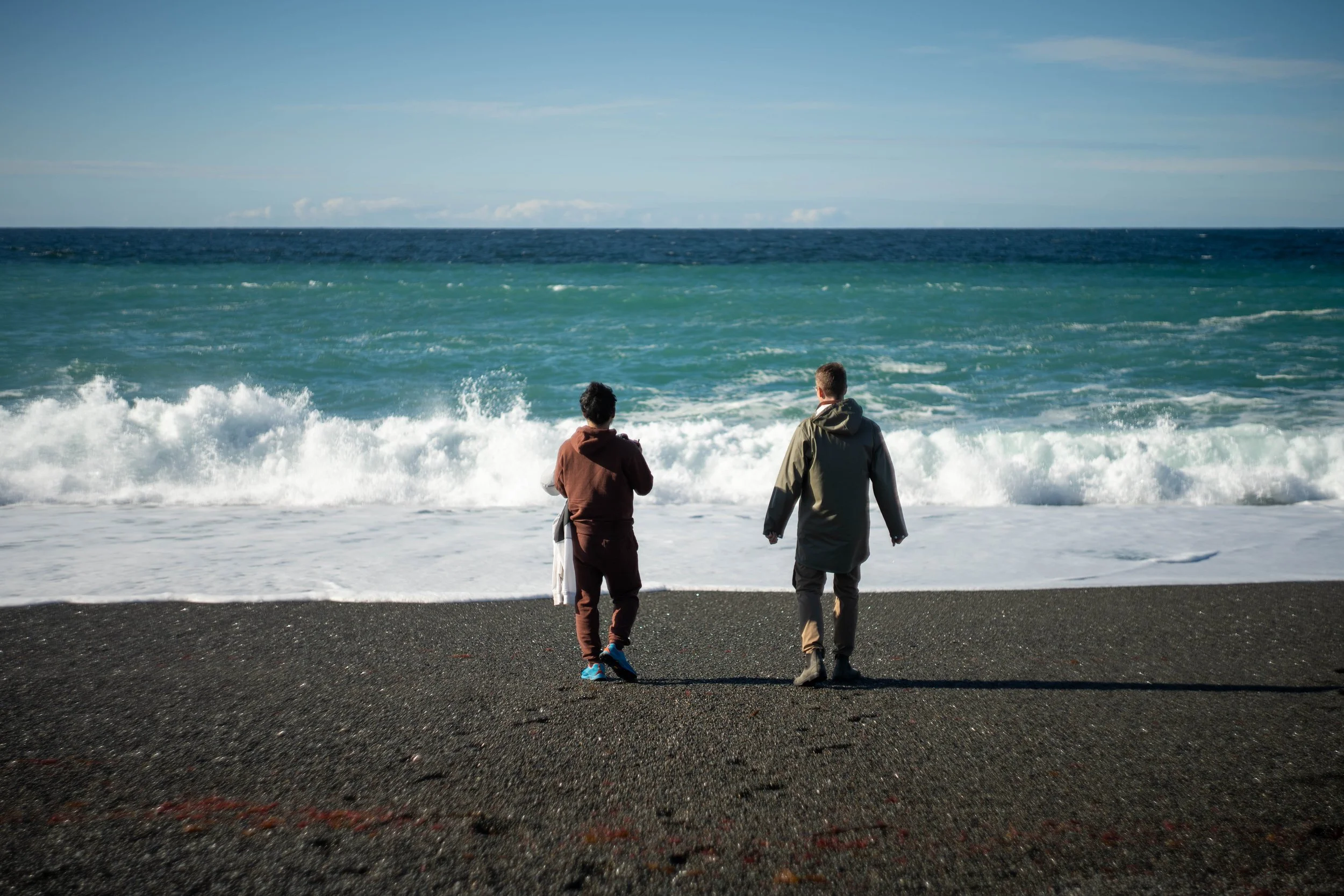 We see two people walking down a stony beach towards some waves.