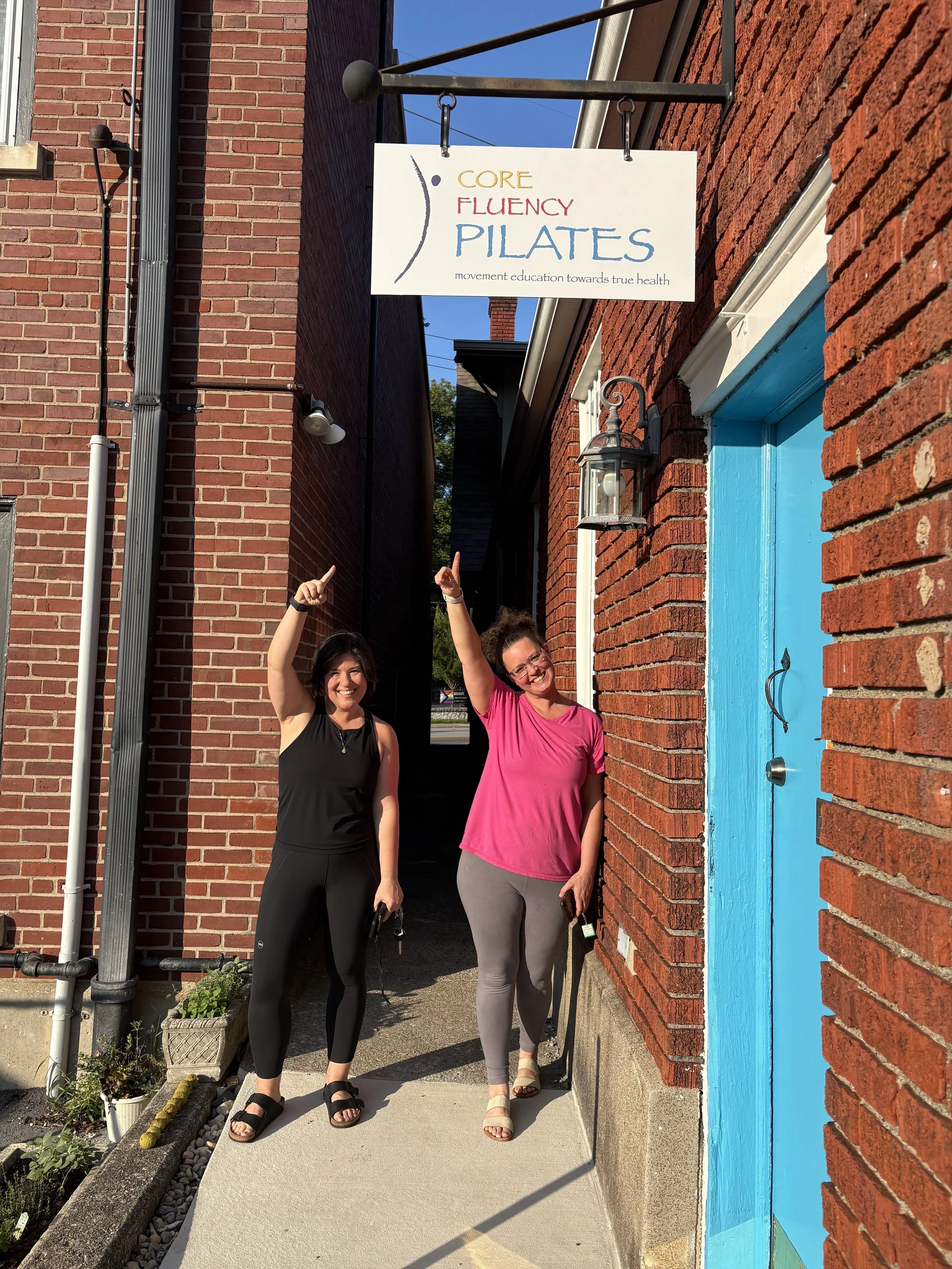 Two clients pointing to Core Fluency Pilates sign at the side studio entrance