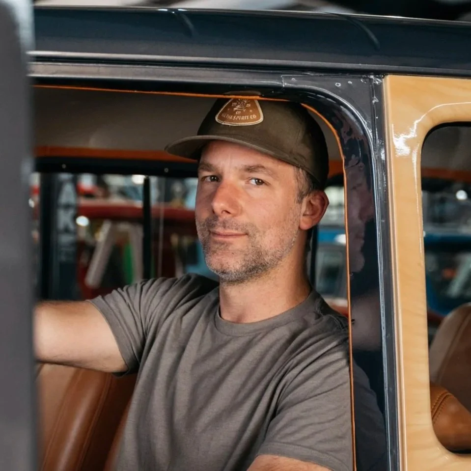 Man with a beard wearing a cap and a gray t-shirt sitting in the driver's seat of a vehicle.