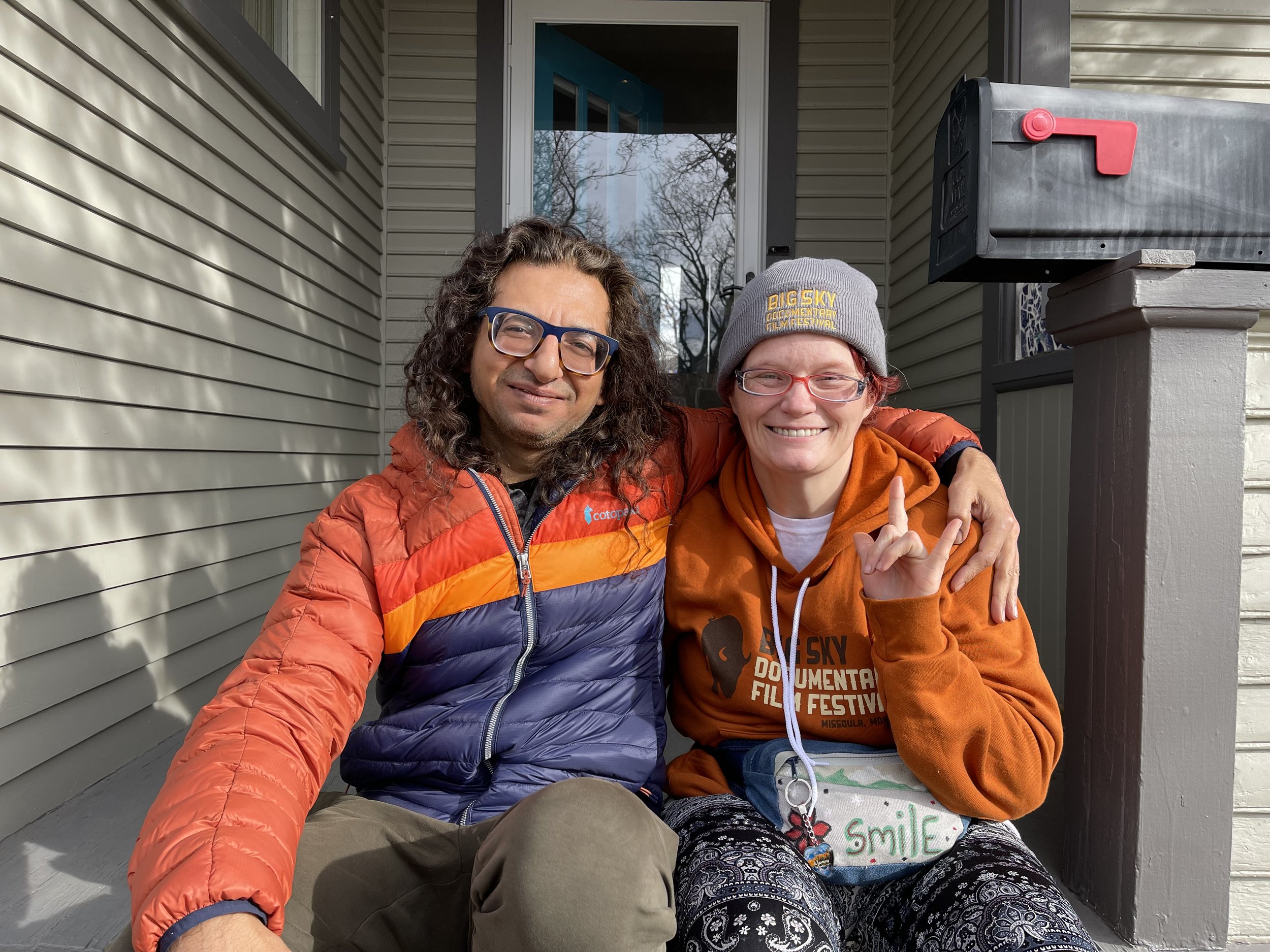 Two smiling people sitting outdoors on a porch. The person on the left has curly hair, glasses, and is wearing a red, orange, and purple jacket. The person on the right has short hair, glasses, and is wearing a gray beanie, orange hoodie with text, a