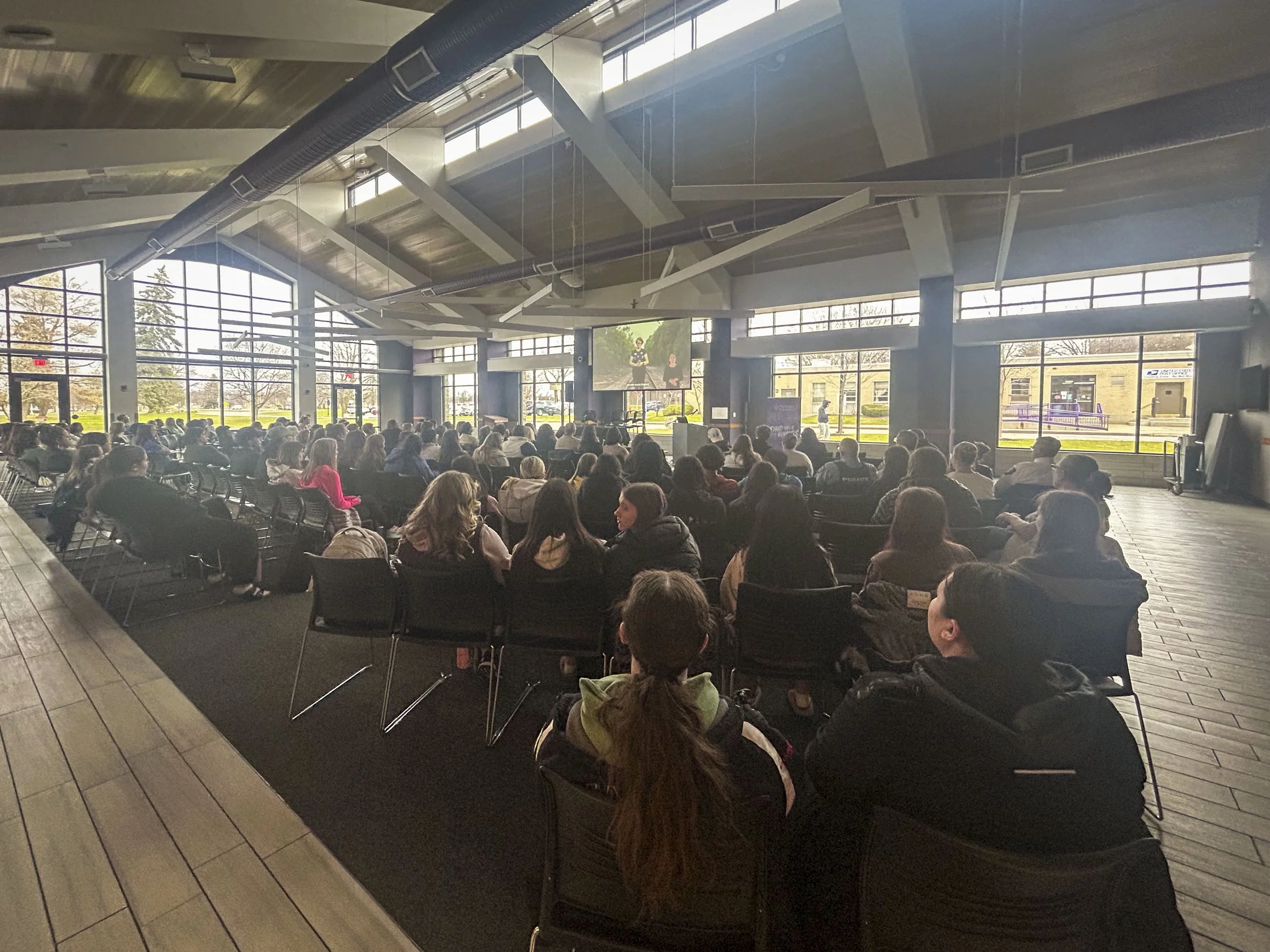 Large indoor conference or event space filled with seated audience facing a stage or presentation area. The room has large windows on two sides, allowing natural light, and a large screen or monitor is positioned at the front displaying two speakers.