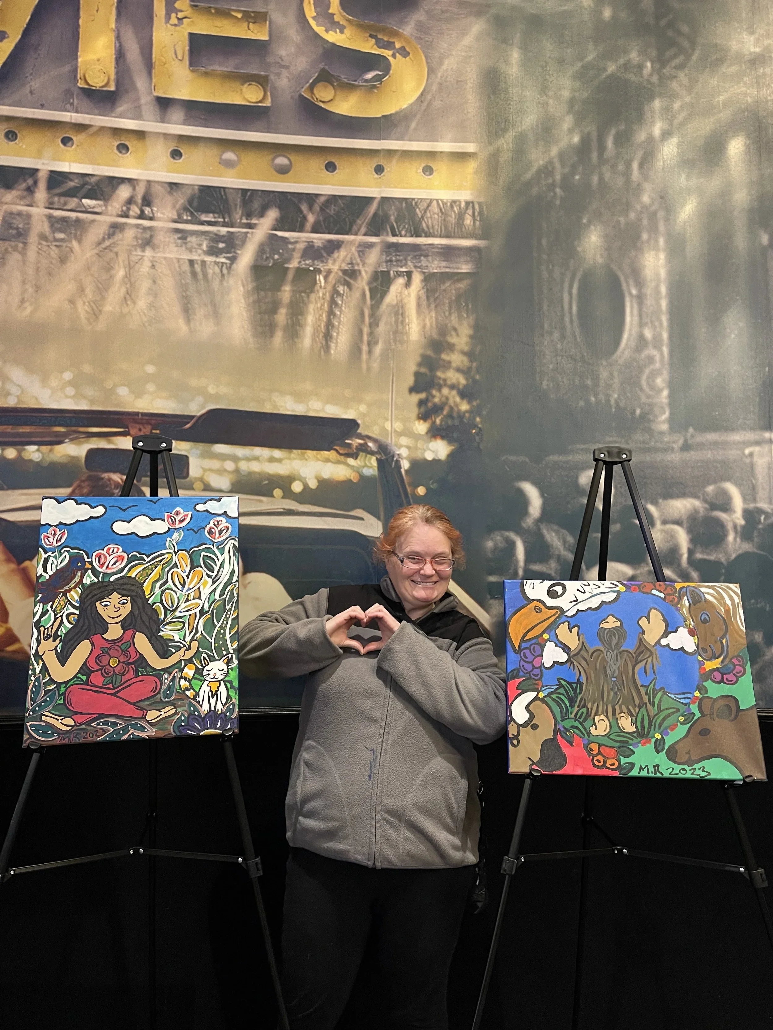 A woman with glasses and a gray jacket smiling and making a heart shape with her hands in front of a black background, flanked by two colorful paintings on easels. The paintings feature vibrant, abstract, and nature-inspired art with floral and animal motifs. Behind her, a large mural depicts a vintage car with a yellow sign reading 'YES' and a landscape scene with faint clouds.