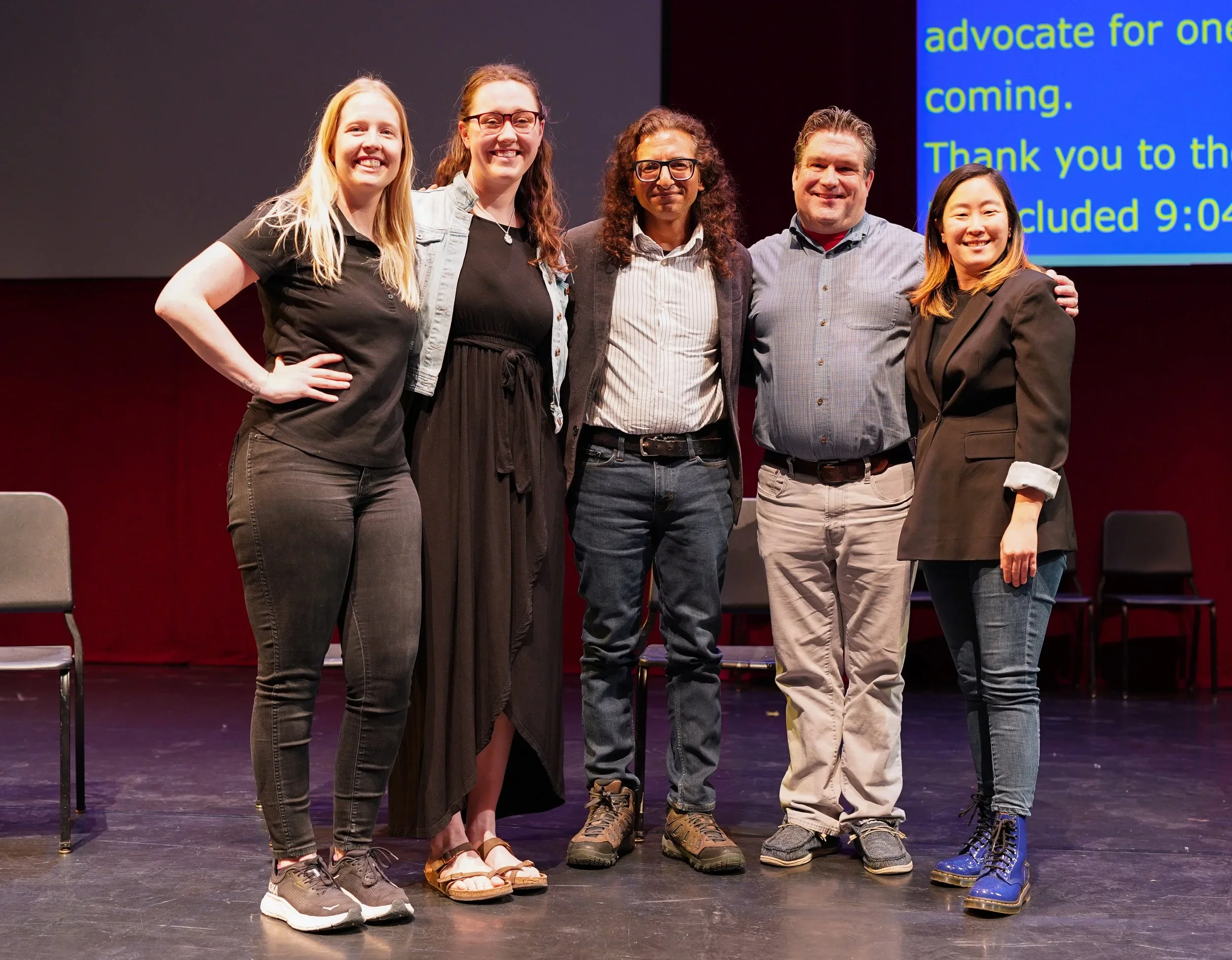 Group of five people standing on stage, smiling, with chairs behind them and a blue screen displaying text in the background.