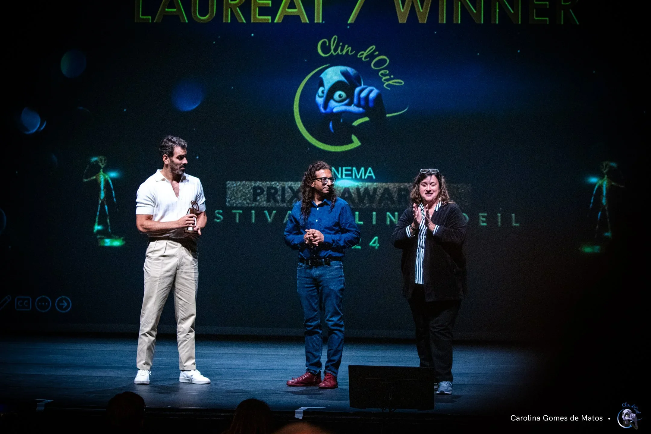 Three people standing on stage during an awards ceremony. The person on the left holds an award. A large screen behind shows a logo with a cartoon character and text. The scene appears to be at the Laureate / Winner event for Clin d'Oeil, with some a