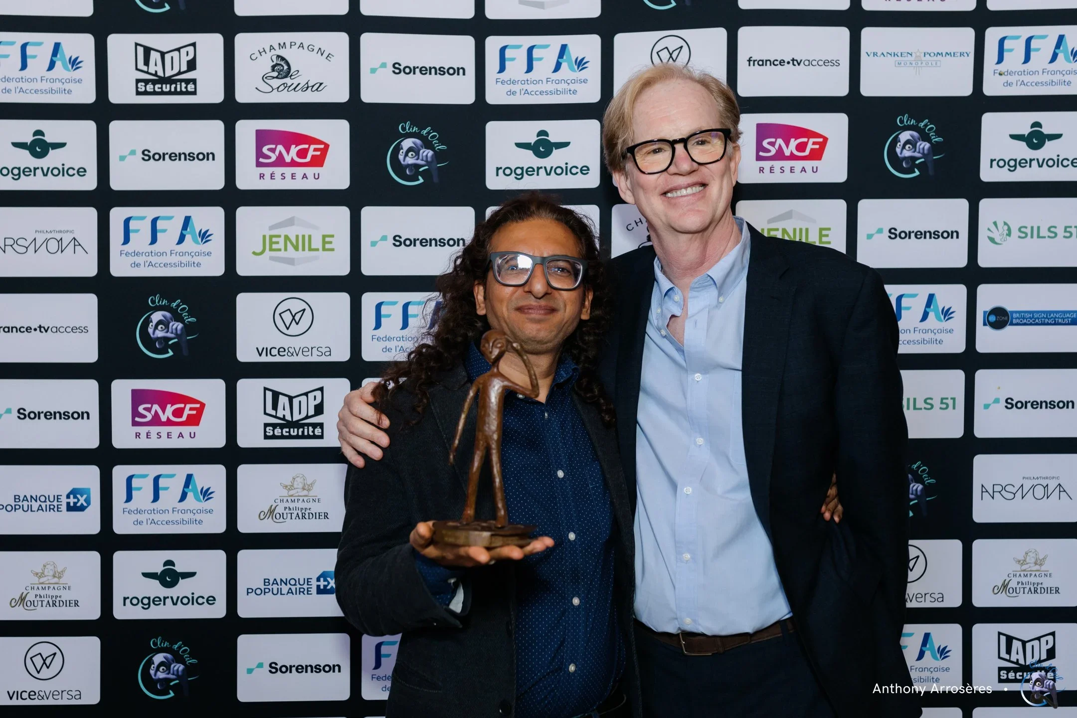 Two men posing at an event with a backdrop covered in logos. The man on the left is holding a trophy and has dark curly hair, glasses, and is wearing a dark blazer. The man on the right is taller, with light hair, glasses, and a blue shirt with a dar