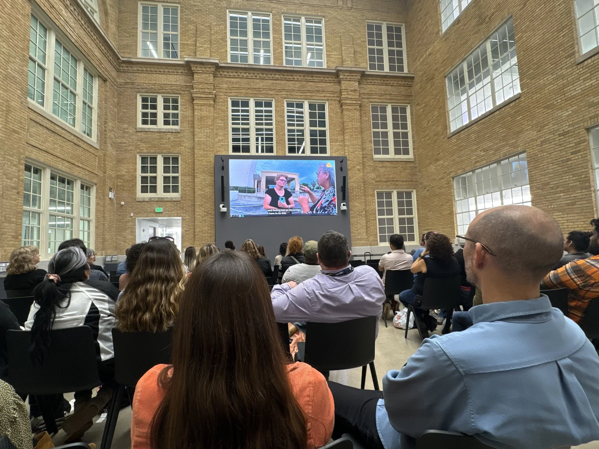 A large group of people seated in chairs watching a presentation in a spacious, multi-story room with numerous large windows and an electronic screen displaying two women in conversation.
