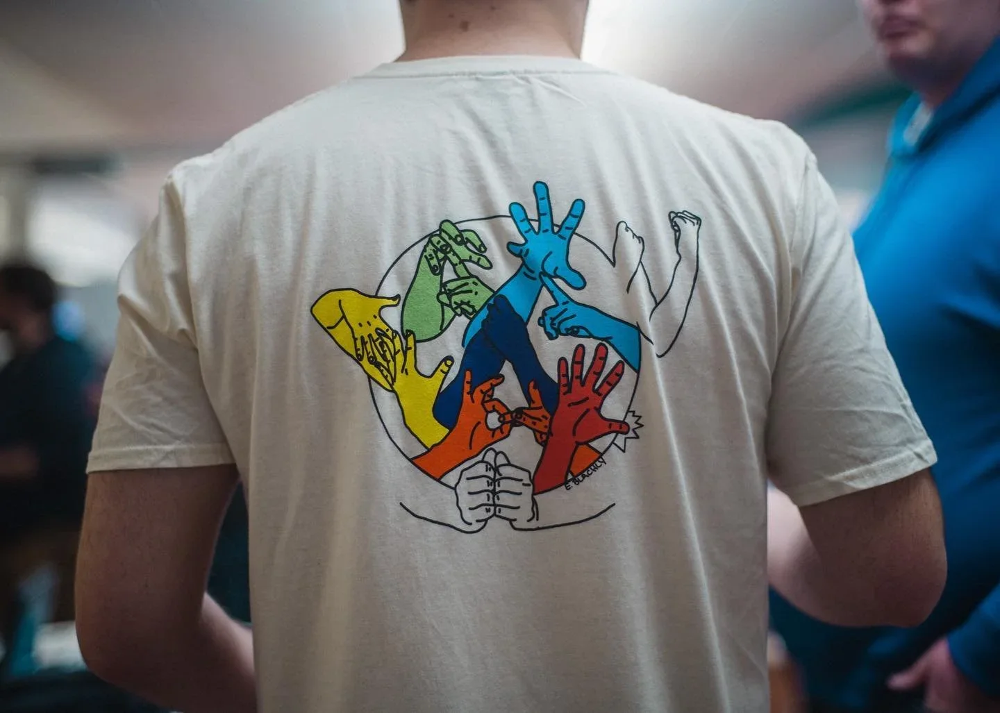 Back of a person wearing a white t-shirt with a colorful illustration of multiple hands in different colors and positions, joining together in a circle.