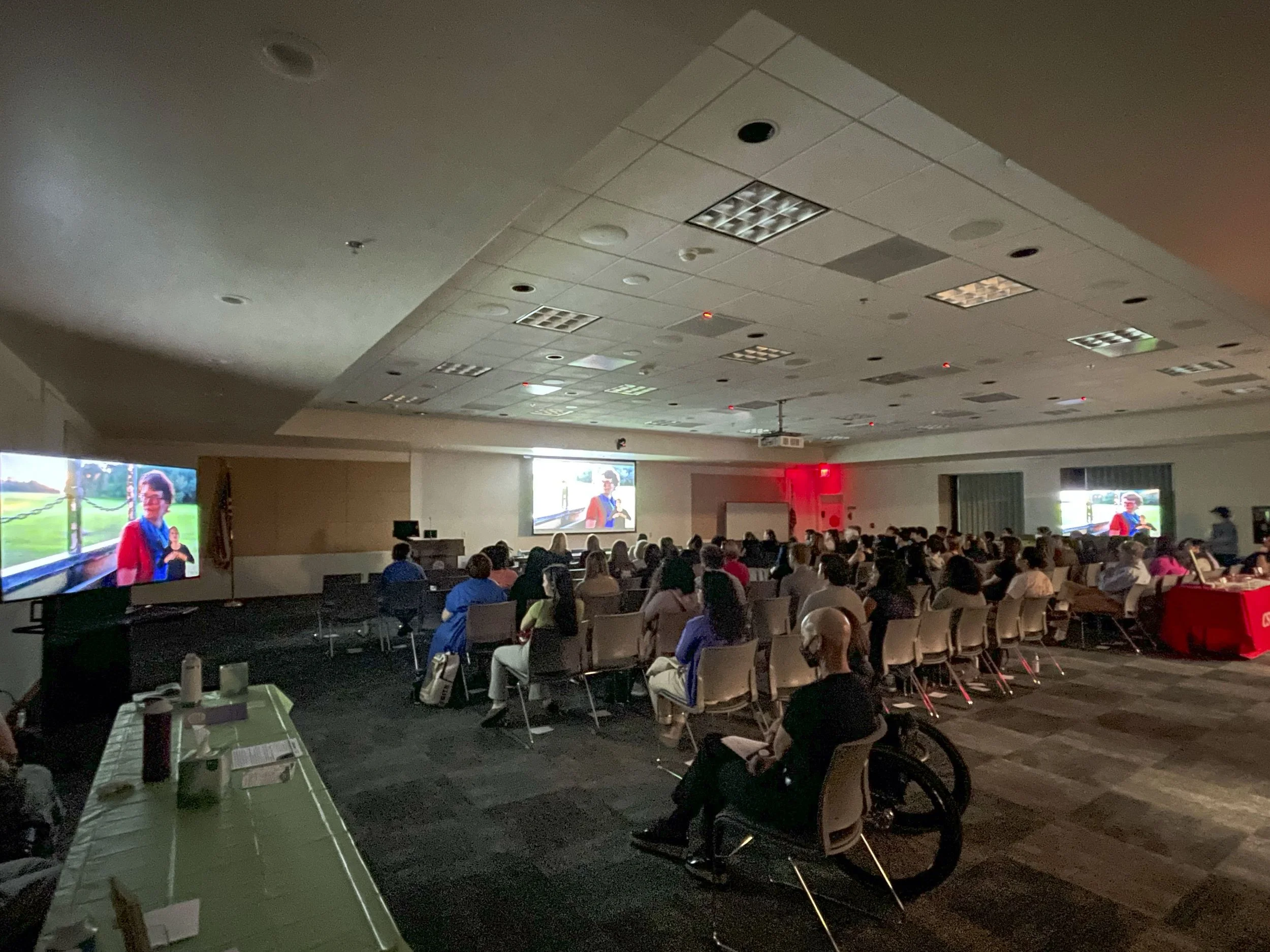 A large group of people seated in chairs in a conference room watching a presentation on multiple screens displaying a woman in a red blazer.