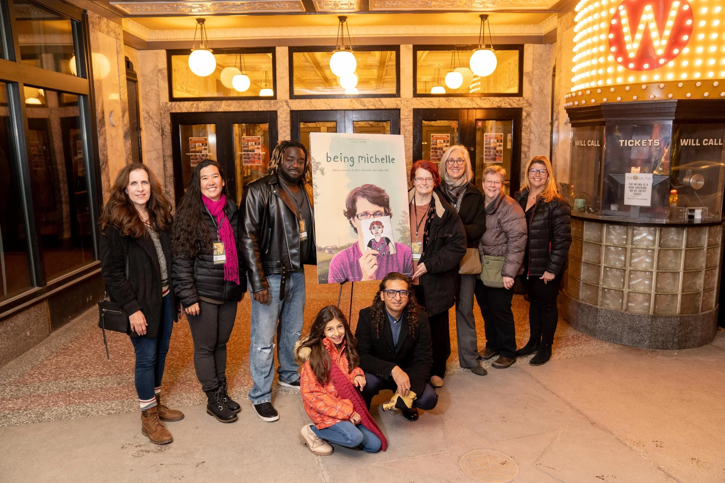 A group of eight adults and two children posing for a photo inside a building with marble walls and illuminated signs. They are standing around a poster that features an illustration of a woman with glasses and curly hair. The environment has warm li