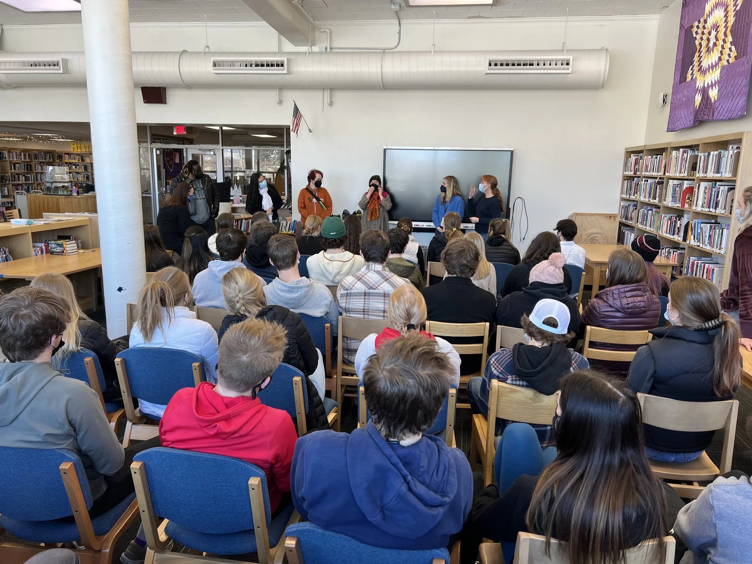 A group of students gathered in a library classroom listening to a presentation, with some standing near a whiteboard and others seated facing forward, wearing masks.