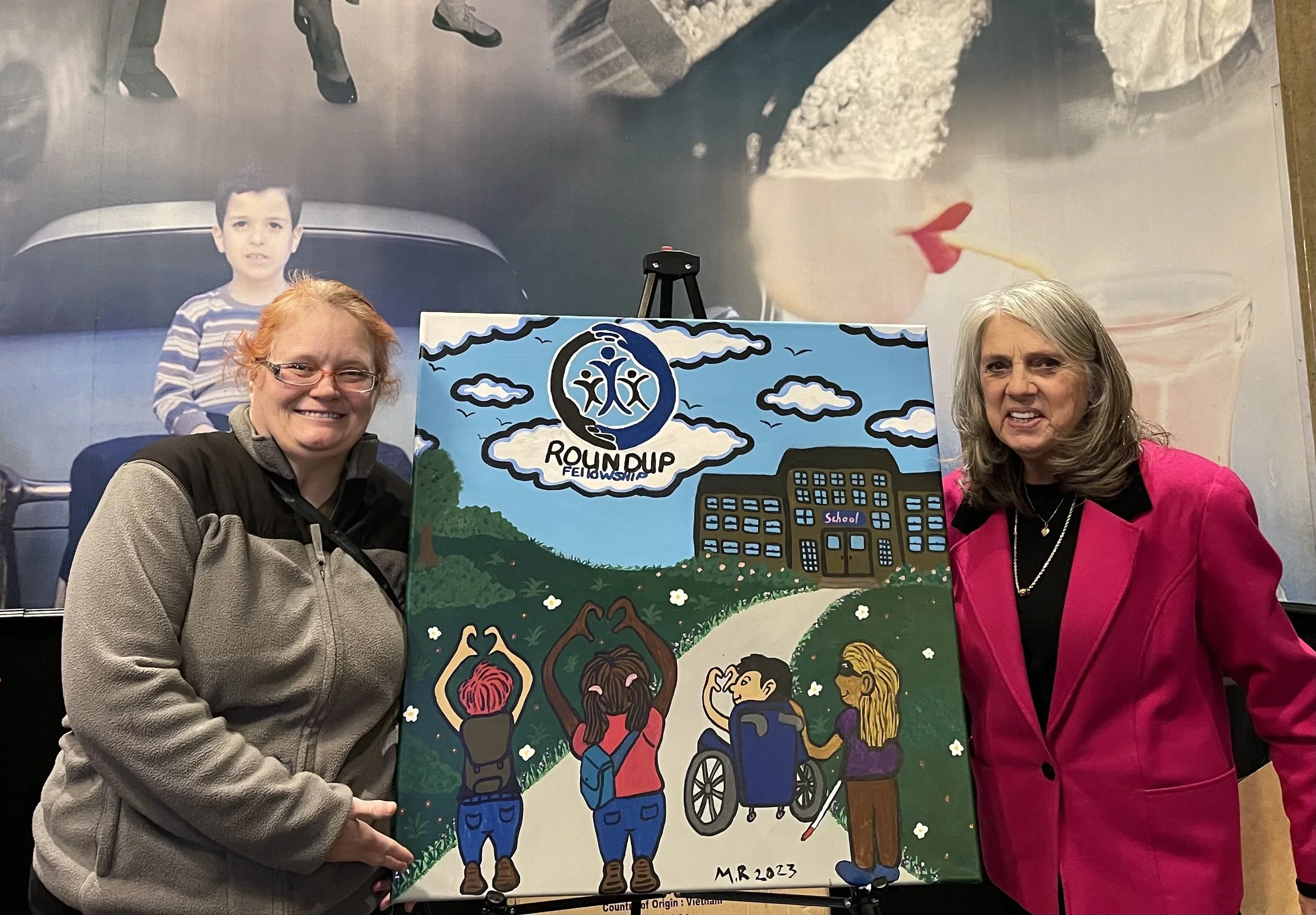 Two women standing in front of a colorful mural holding a sign that reads "Roundup Fellowship". The mural features a large face of a woman with a red lipstick and a lollipop, and a children’s drawing of students walking toward a school building, with mountains and clouds in the background.