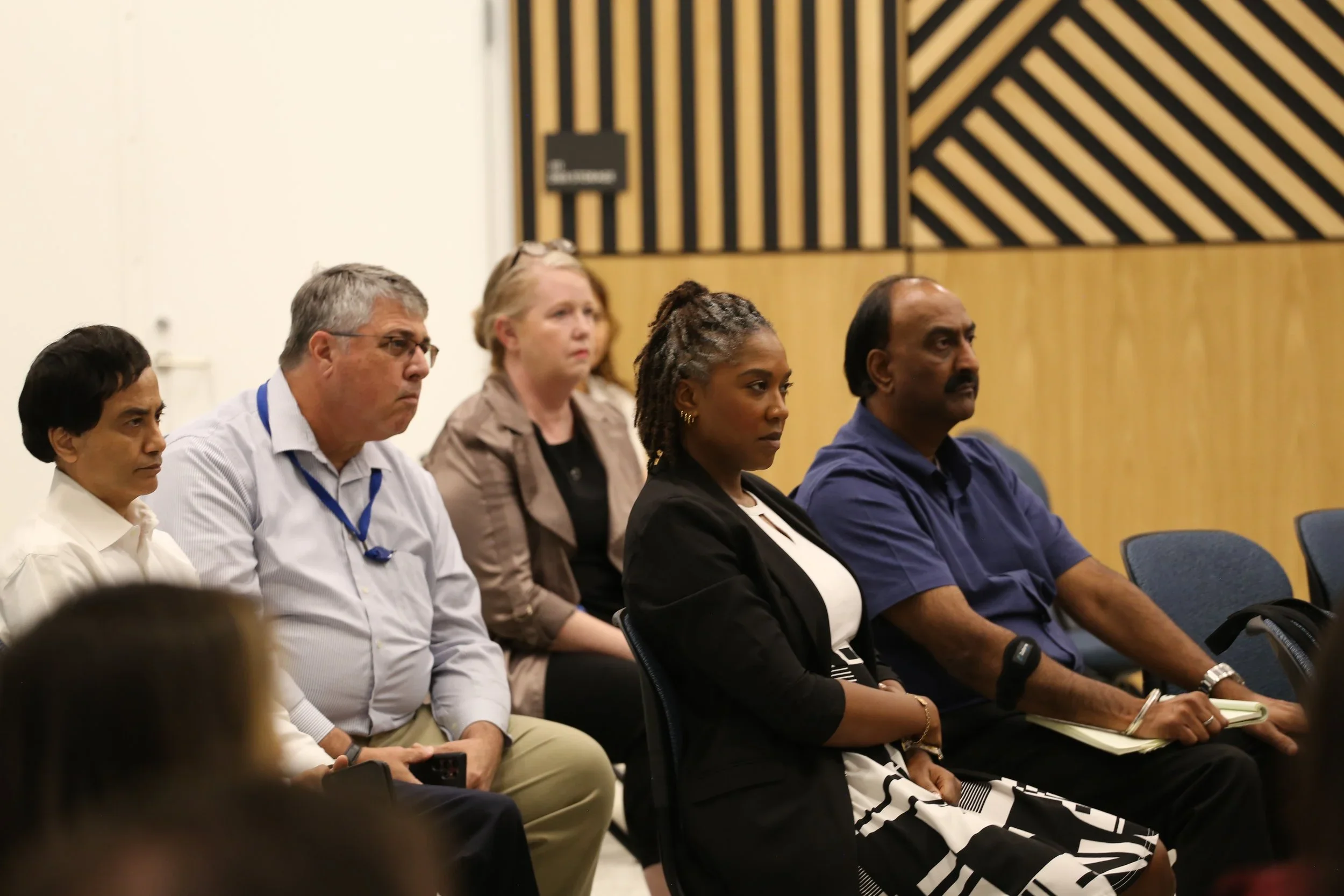 People sitting in a row at a conference or meeting room, attentively listening, with a wooden panel background.