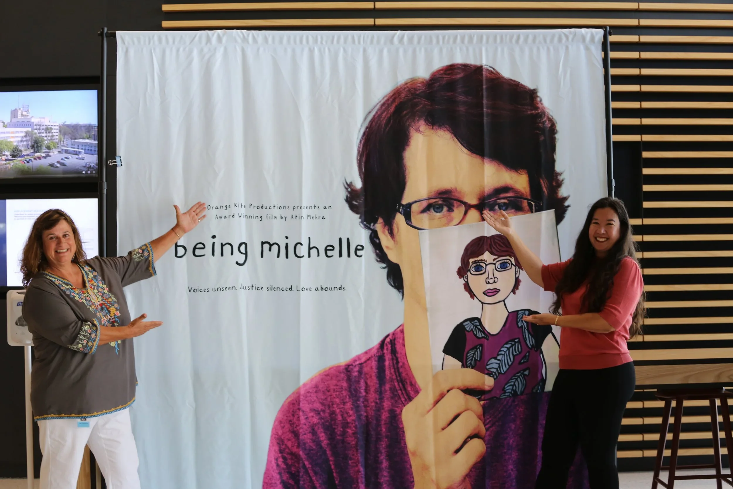 Two women standing in front of a large poster of a woman with short dark hair, glasses, and a purple sweater. The women are smiling and pointing at the poster.