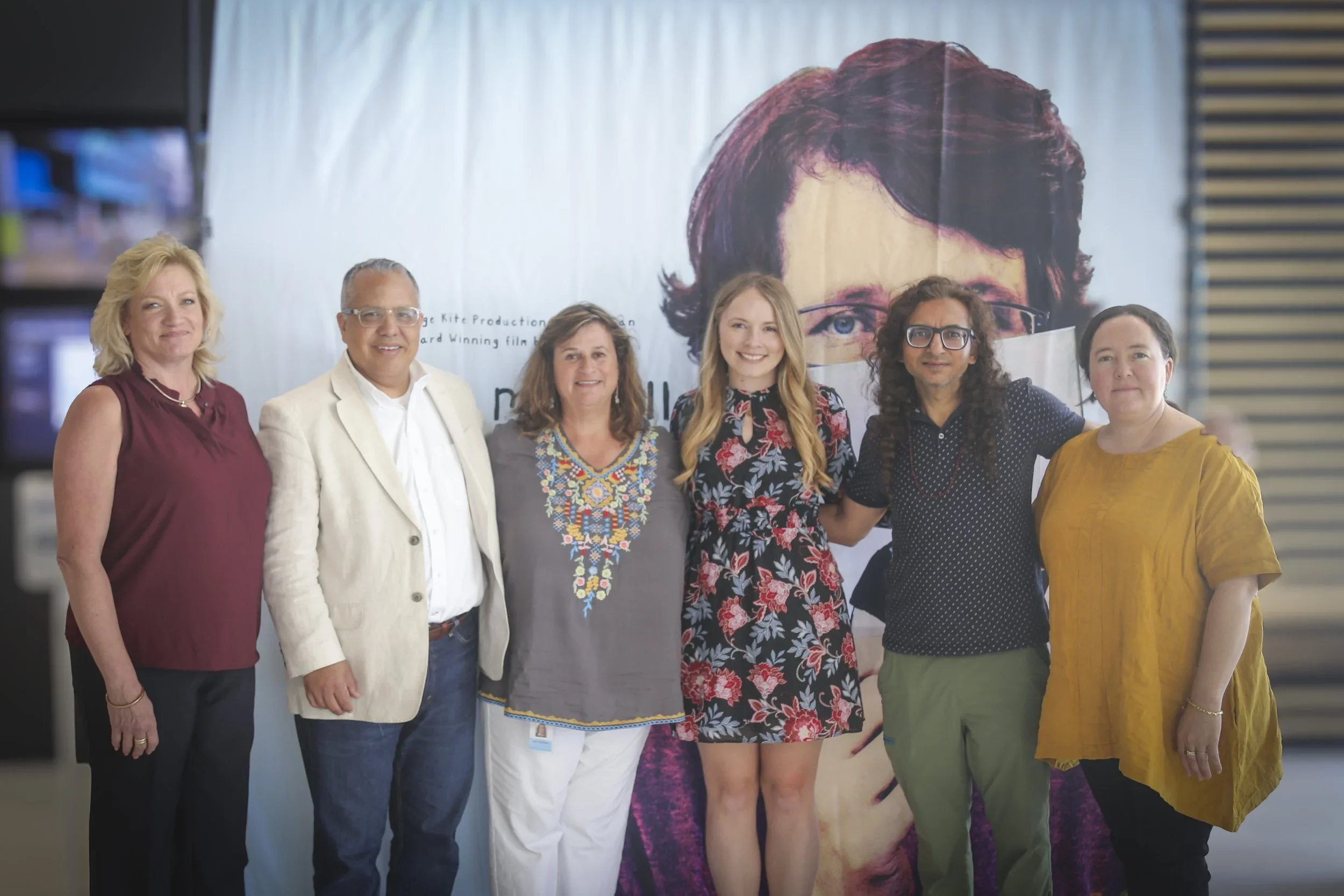Group of six diverse people standing together indoors, smiling, in front of a large poster with a person's face on it.