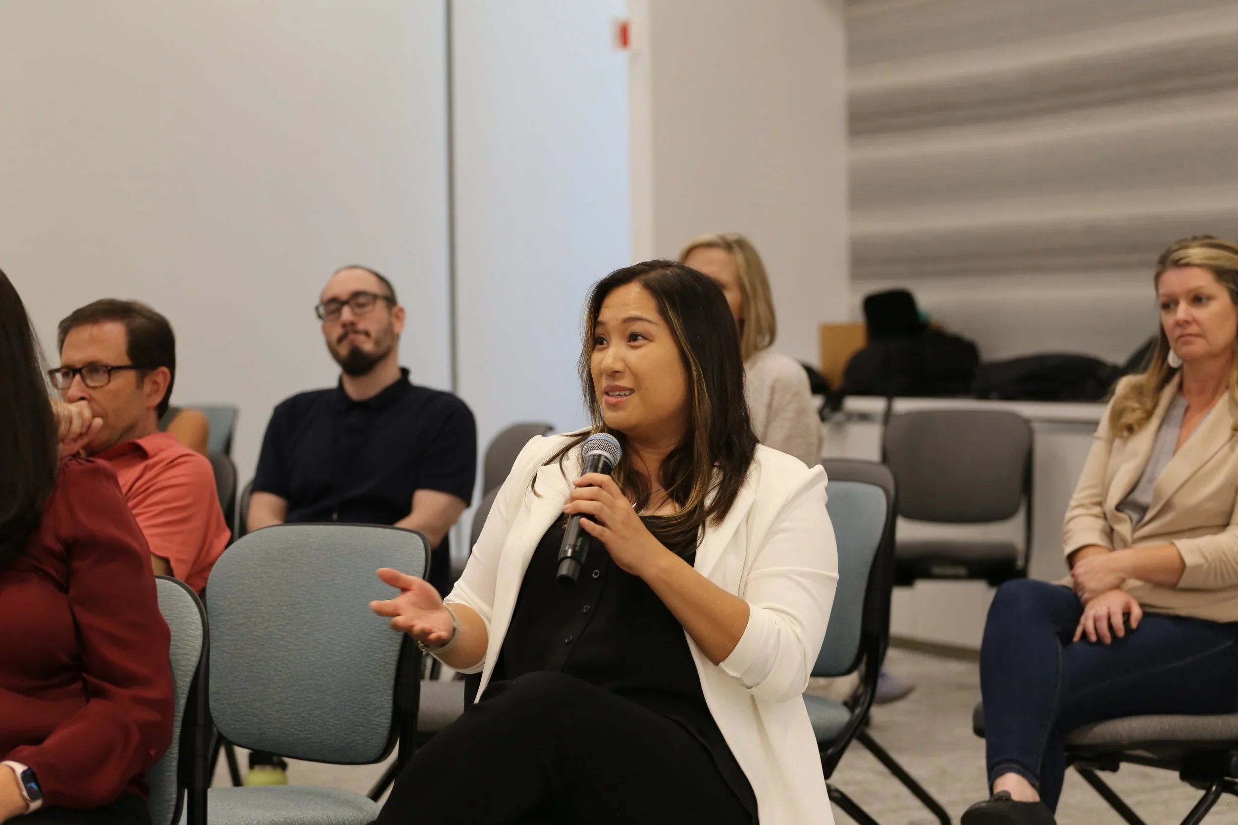 A woman in a white blazer and black top is holding a microphone and speaking during a meeting or conference, sitting among other attendees in a room with gray chairs and neutral walls.