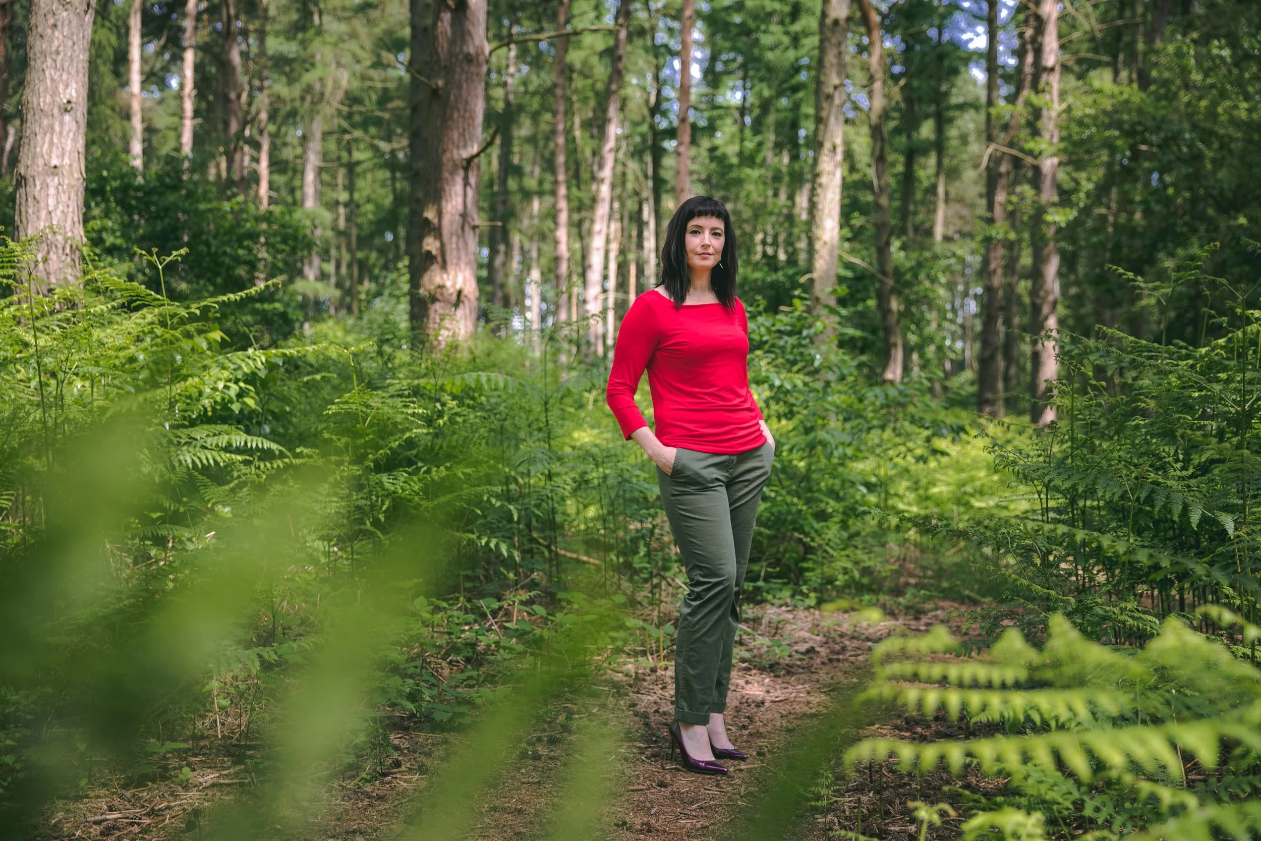 Anne Bryant in a forest with a red top on - Myalgic Encephalomyelitis Practitioner