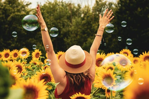 A woman in a panama hat joyfully throws her arms up as she stands in a sunflower field amongst bubbles