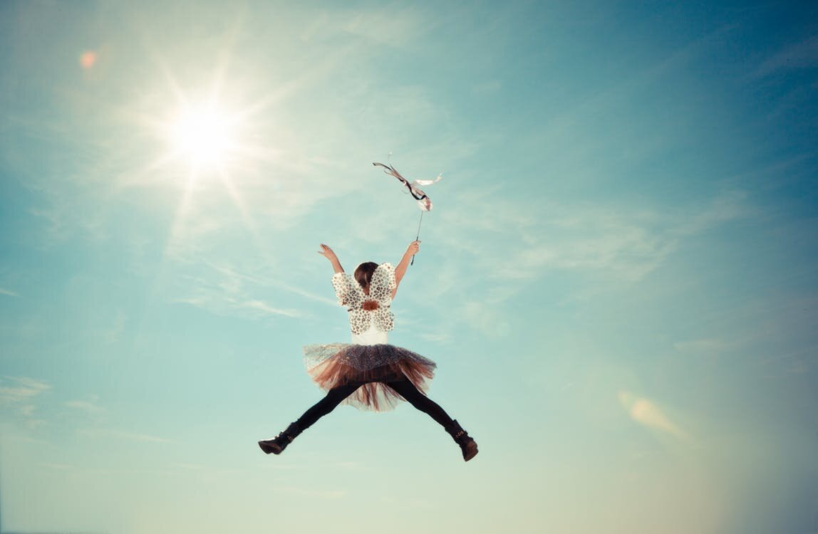 a woman in wings and tutu jumps for joy in a sunny sky