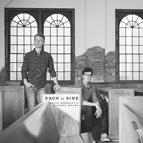Two young men in a church with wooden pews and large arched windows. One is standing with hands in pockets, the other is sitting and leaning on the pew. There is a sign that says "Each by Side" with the names David Benedict & Michael Moore.