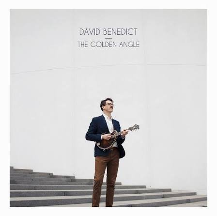 A man in a navy jacket and brown pants playing a ukulele or guitar in front of a white wall with the text 'David Benedict The Golden Angle'.