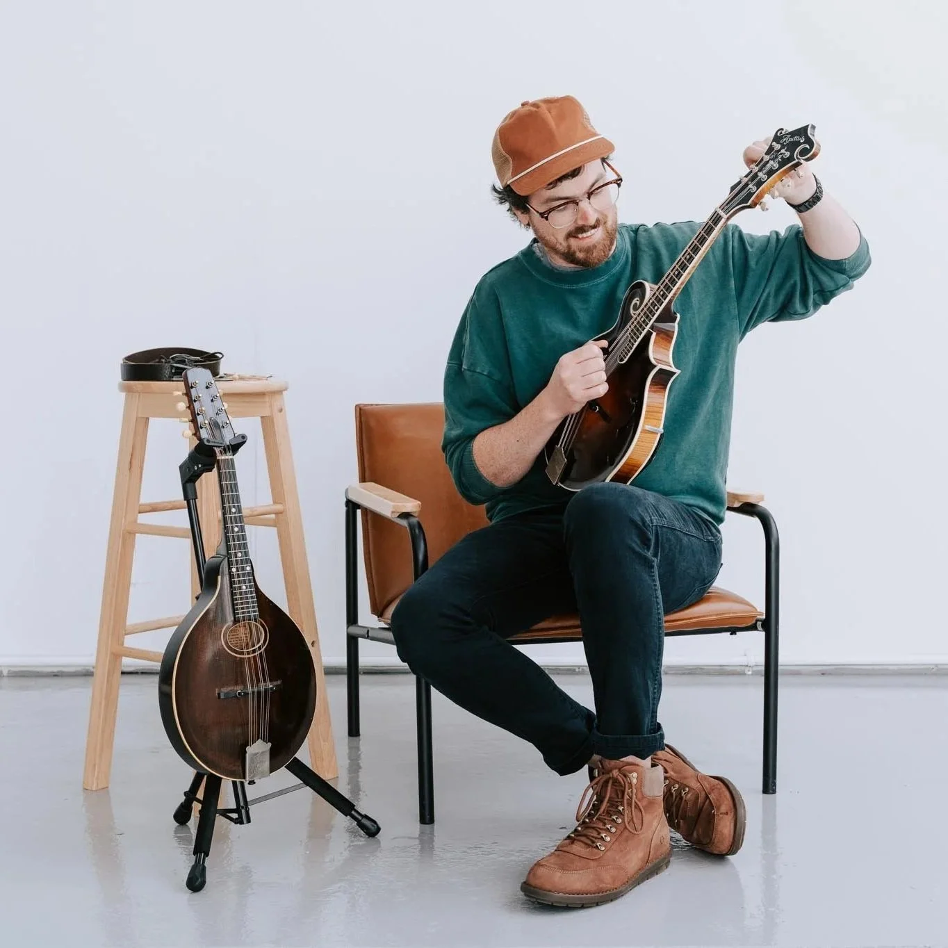 A man with glasses, a beard, and brown boots sitting on a chair, playing a mandolin. There is a guitar on a stand and another mandolin on a stool beside him, with a plain white wall background.
