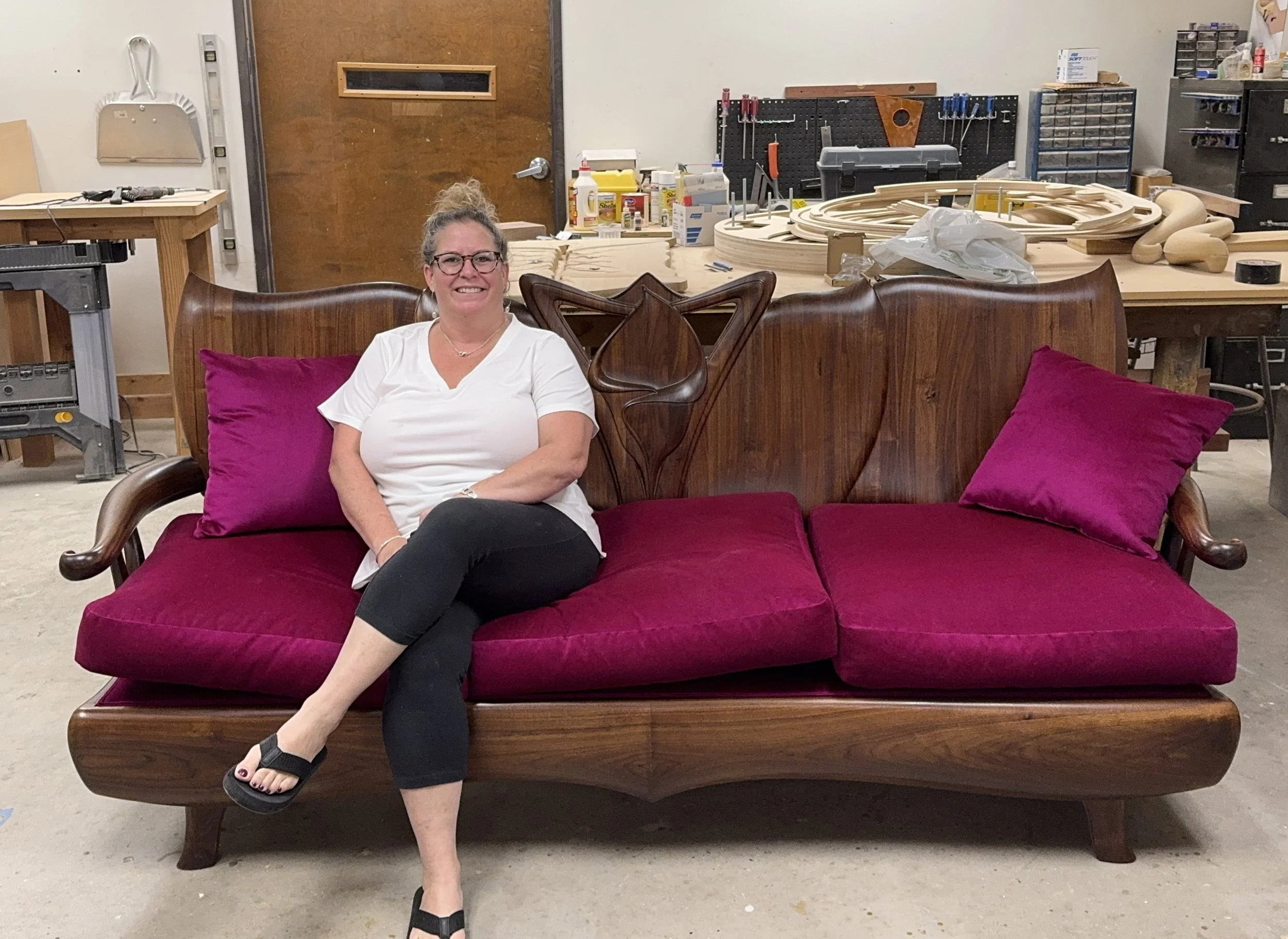 A woman with glasses, wearing a white shirt and black pants, sitting on a walnut sofa with fuschia cushions and pillows in a woodworking workshop.