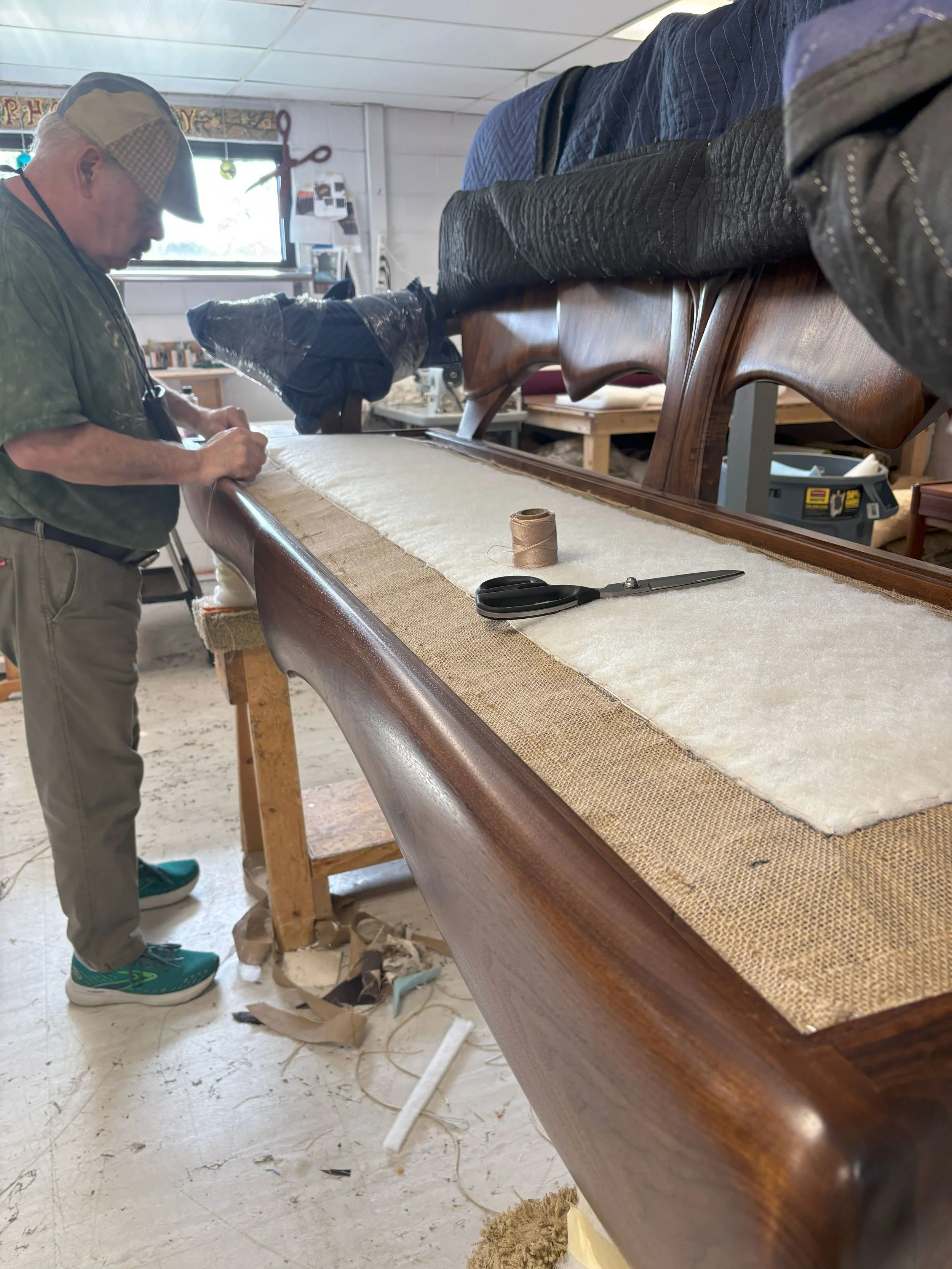 Man working on upholstery of a wooden sofa frame in a workshop, with tools like scissors and thread on the frame.