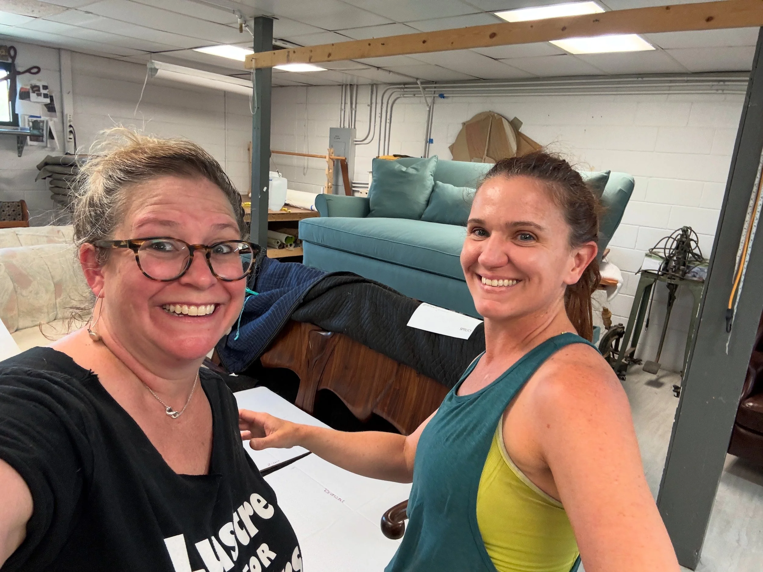 Two women smiling in a warehouse with furniture and tools in the background.