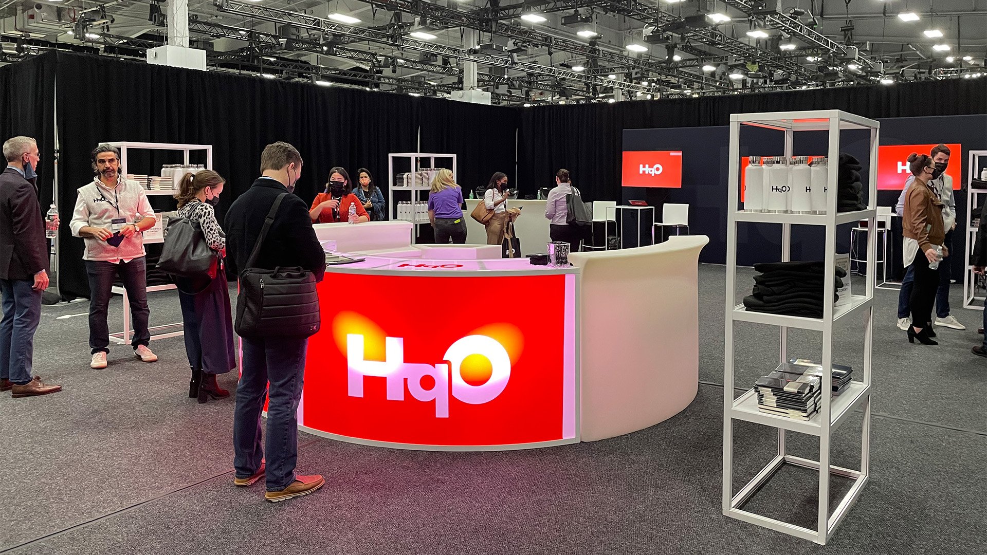 People gather around a trade show booth with a large red and white 'HqO' logo. The booth features a curved counter, shelving units with branded merchandise, and a background with multiple booths and black curtains. Attendees are wearing masks and engaging with the booth.