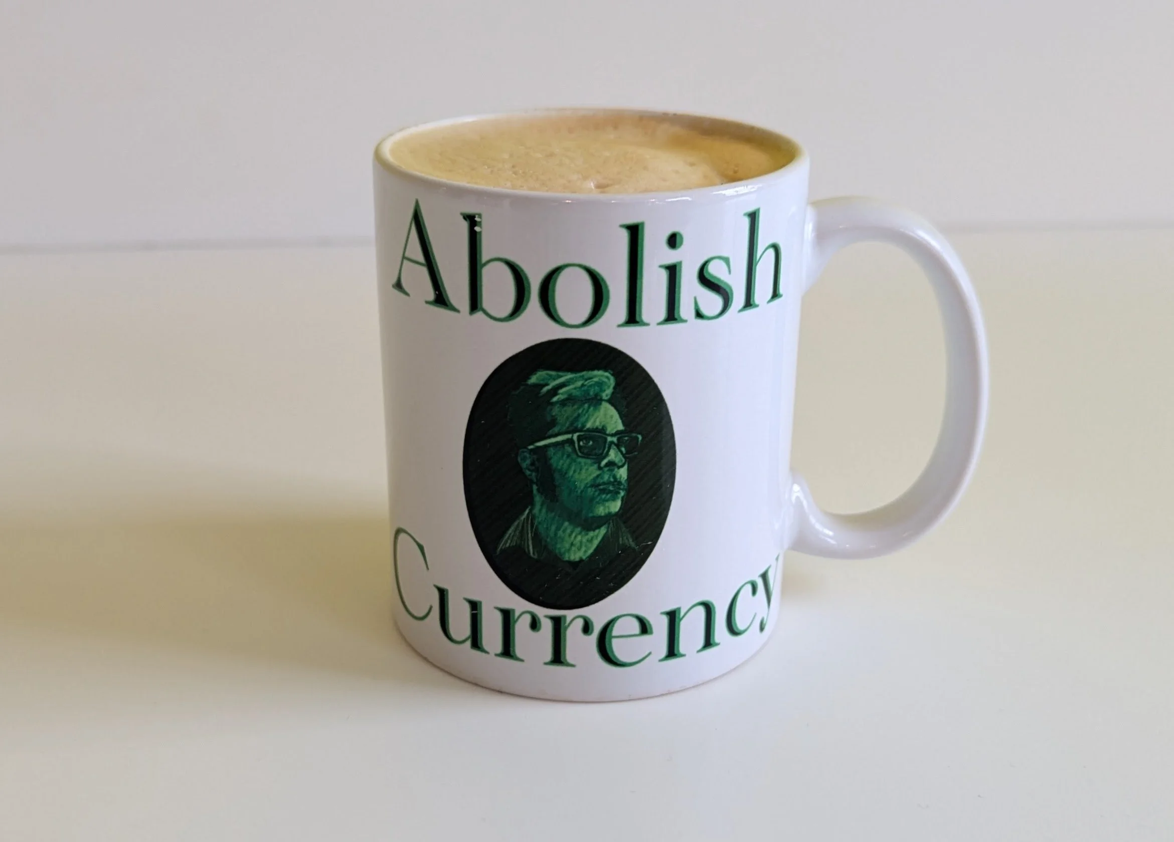 A coffee mug with a hand-drawn portrait of a man with glasses, vaguely in the style of the portraits of presidents on US dollars. Text on the mug reads "Abolish Currency"