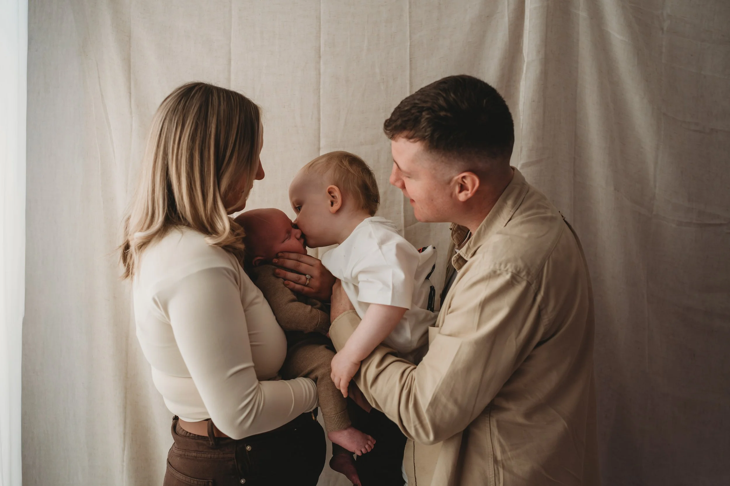 A family of four with two young children, a baby and a toddler, sharing a moment of affection indoors, with neutral-colored curtain background.