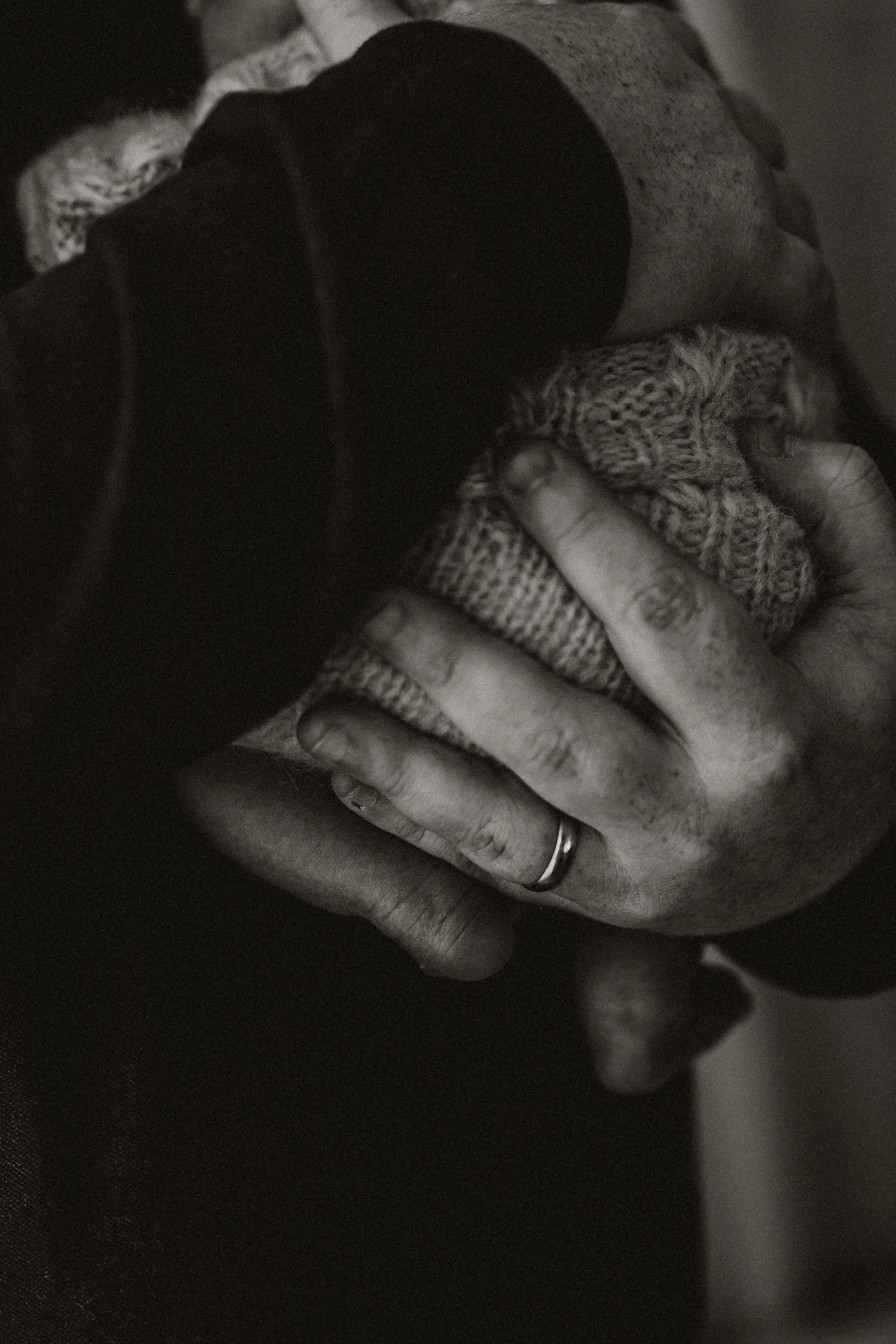 A person's hand with a wedding band on the ring finger, gently holding and pressing against their face, which is shrouded in shadow. The person is wearing a patterned knit glove and a dark long-sleeve shirt.