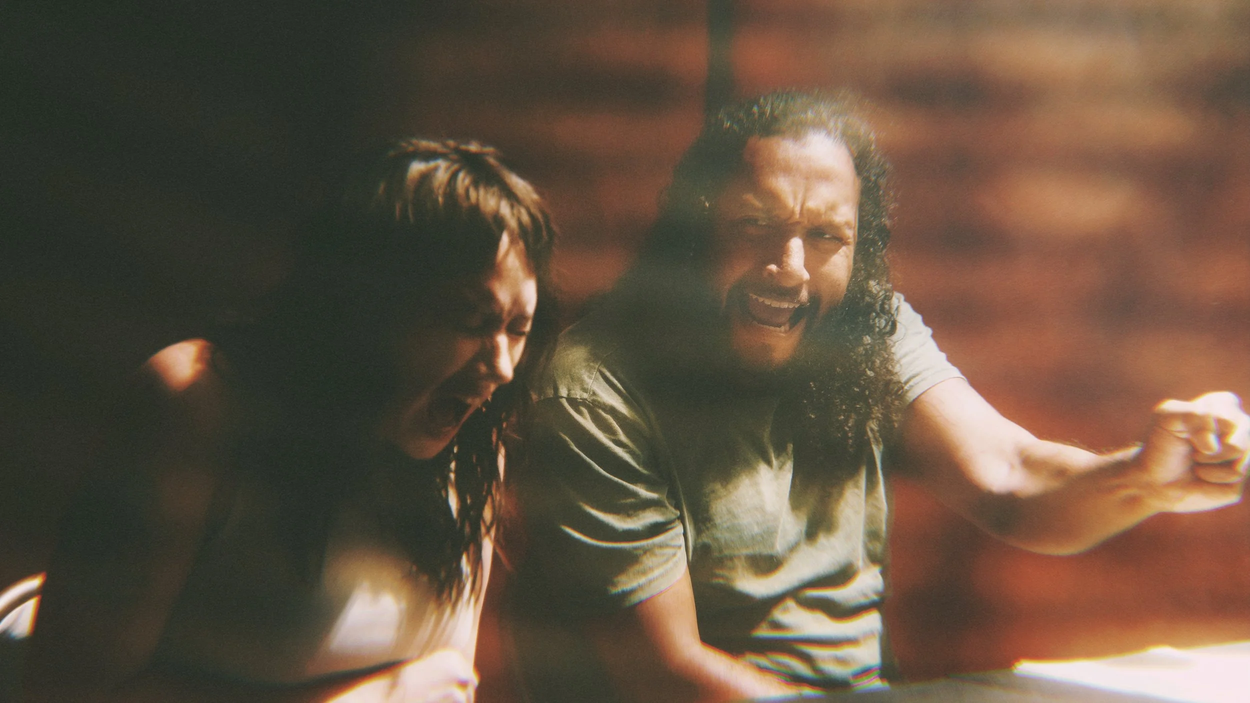 Two men with long hair appear to be in a moment of intense emotion, possibly anger or protest, with clenched fists and open mouths.